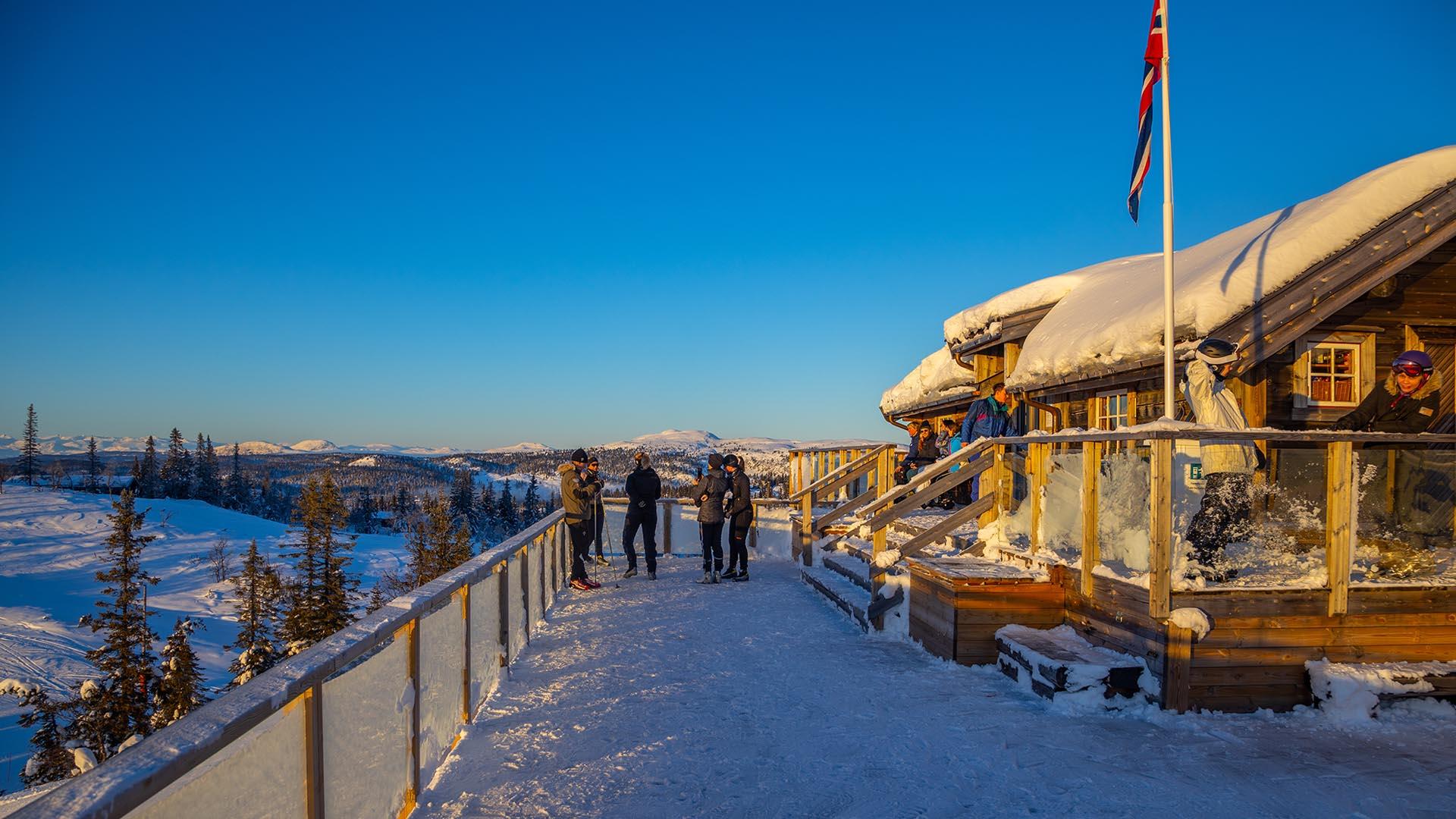 Folk står på verandaen til et serveringssted på toppen av et alpinsenter og nyter en skyfri vinterdag.