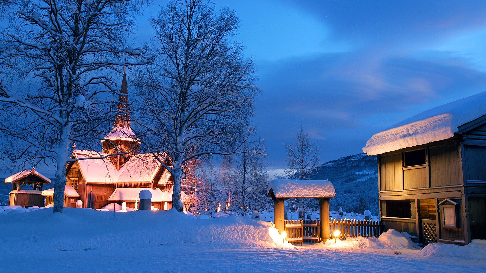A snowy day in the blue hours, a stave church in the background and an old wooden building in the foreground.