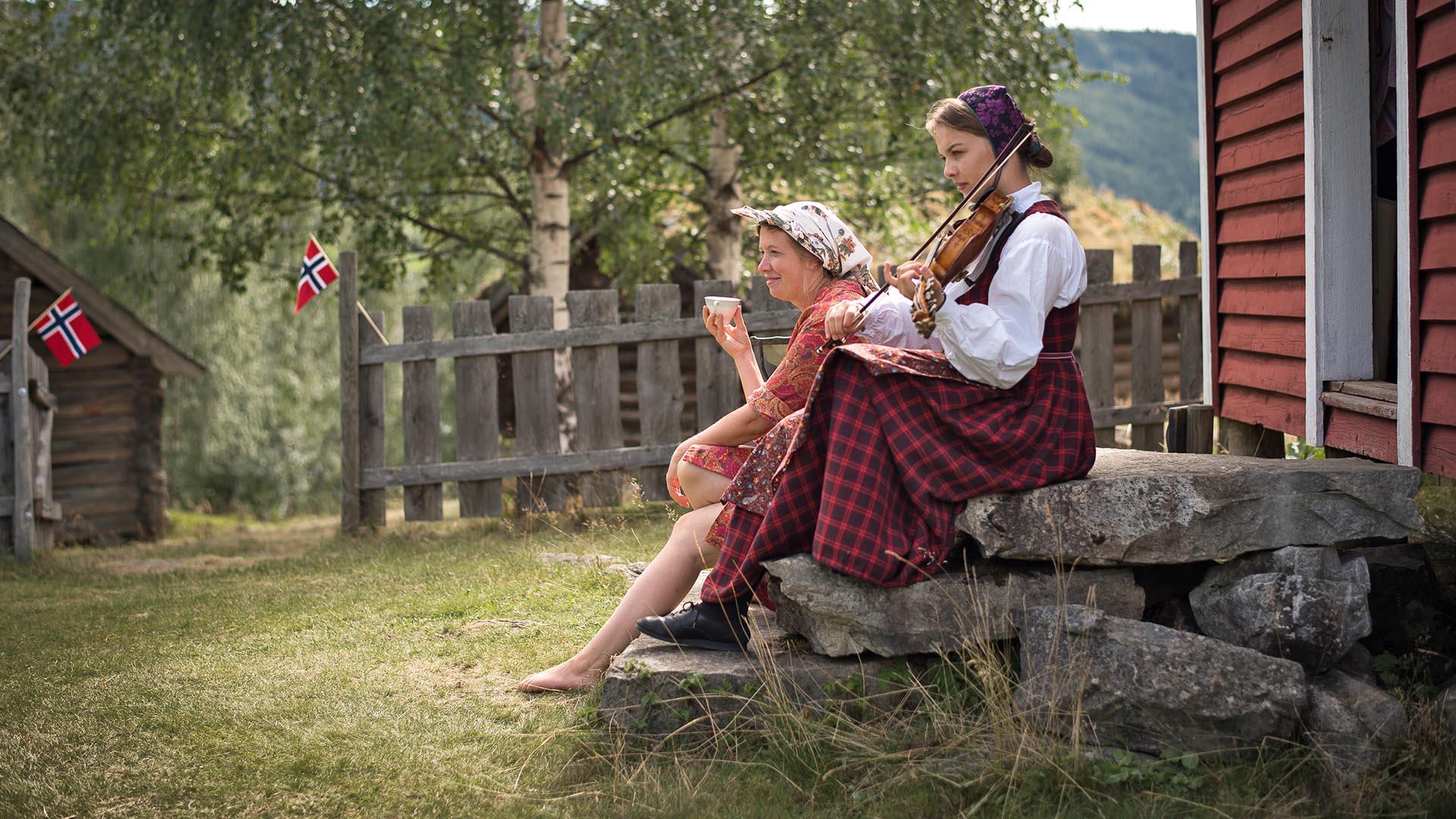 Zwei junge Mädchen in Volkstracht sitzen auf der Steintreppe vor einem alten Haus im Valdres Volksmuseum und genießen den schönen Sommertag. Eine von ihnen spielt auf der Hardangerfiedel.