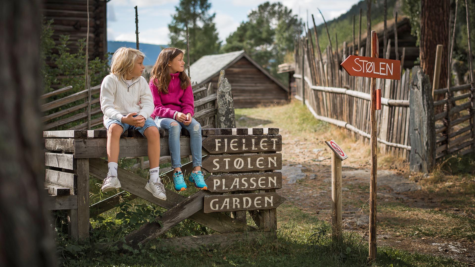 Kinder sitzen auf einem Holzzaun neben Holzschildern, die Besuchern des Valdres Volksmuseums den Weg weisen