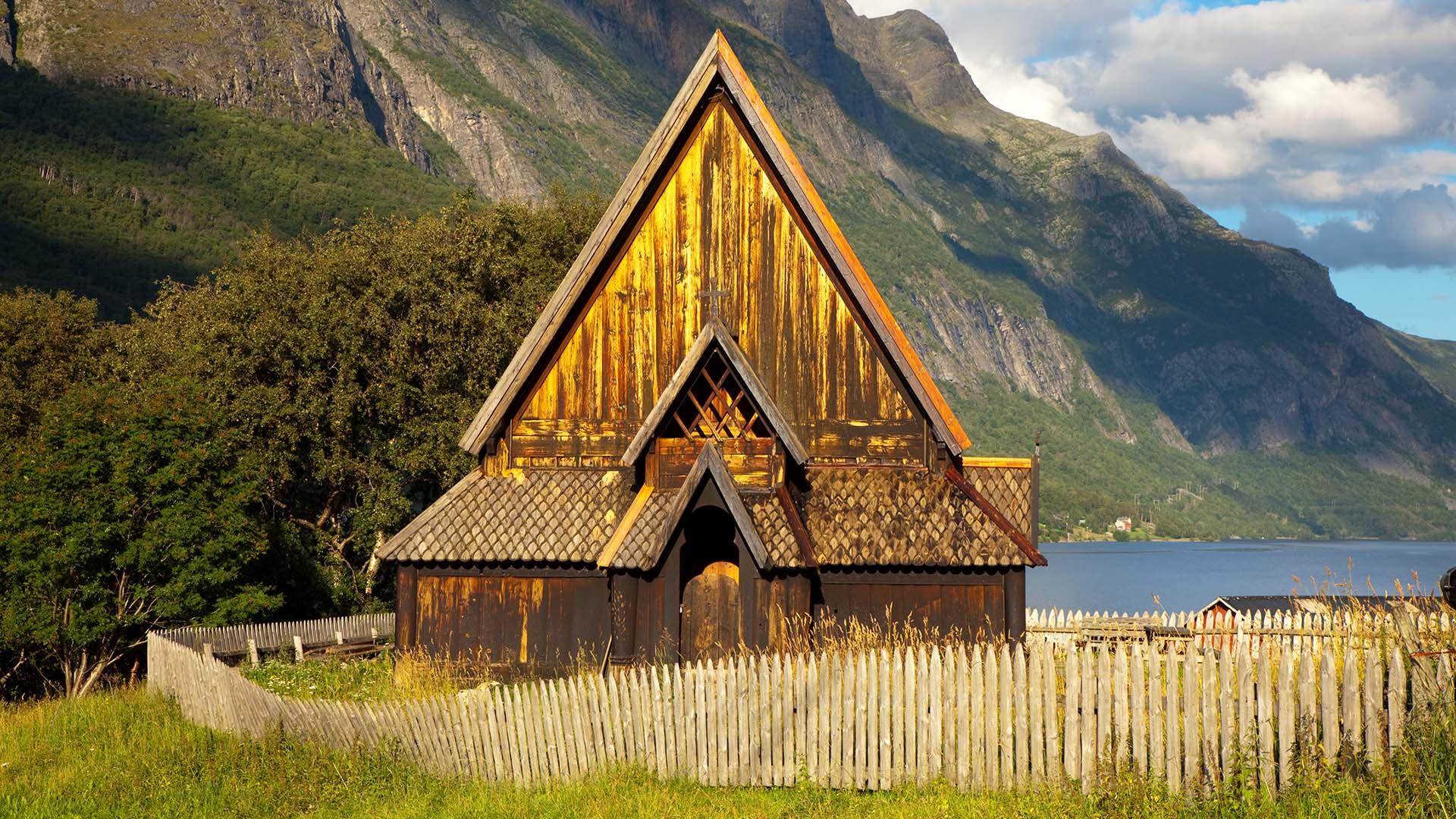 A small wooden stave church with a triangular form fenced in on a green field with a lake and steep hillsides beyond.