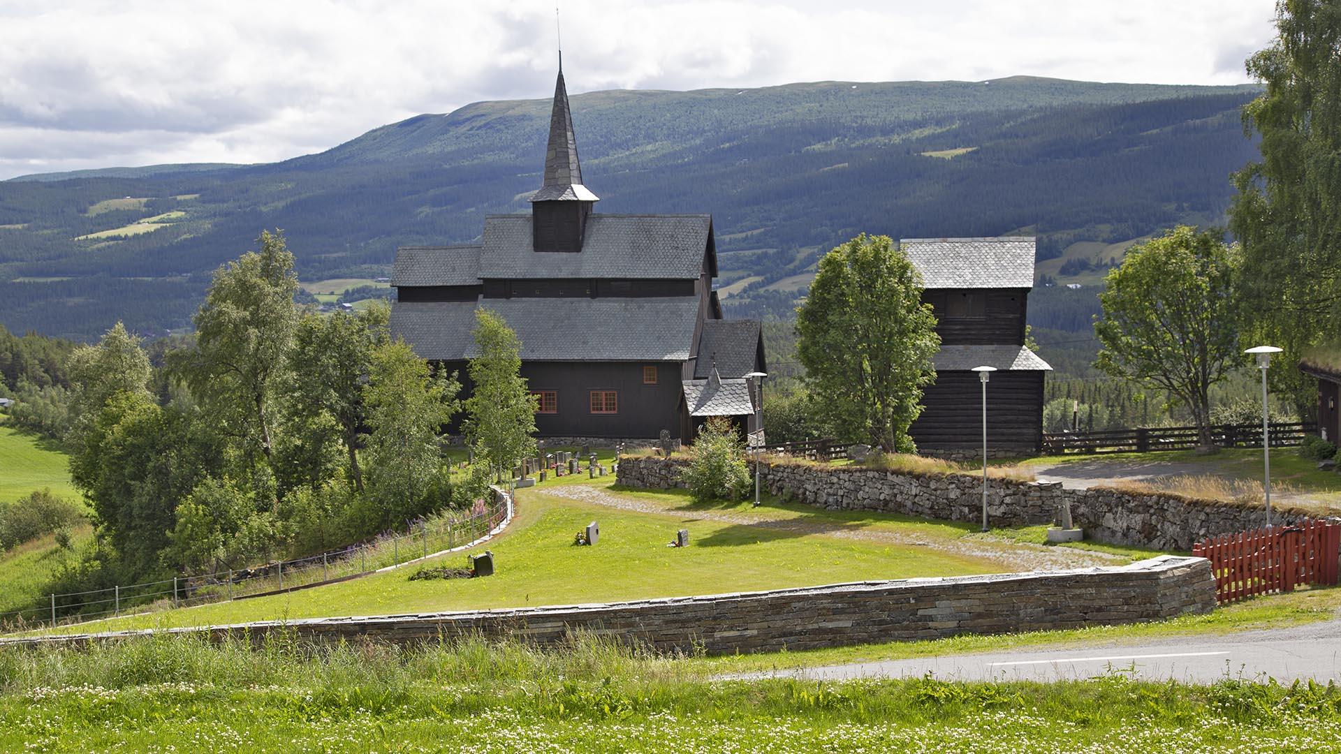 Tar colored stave church surrounded by green trees and grass.