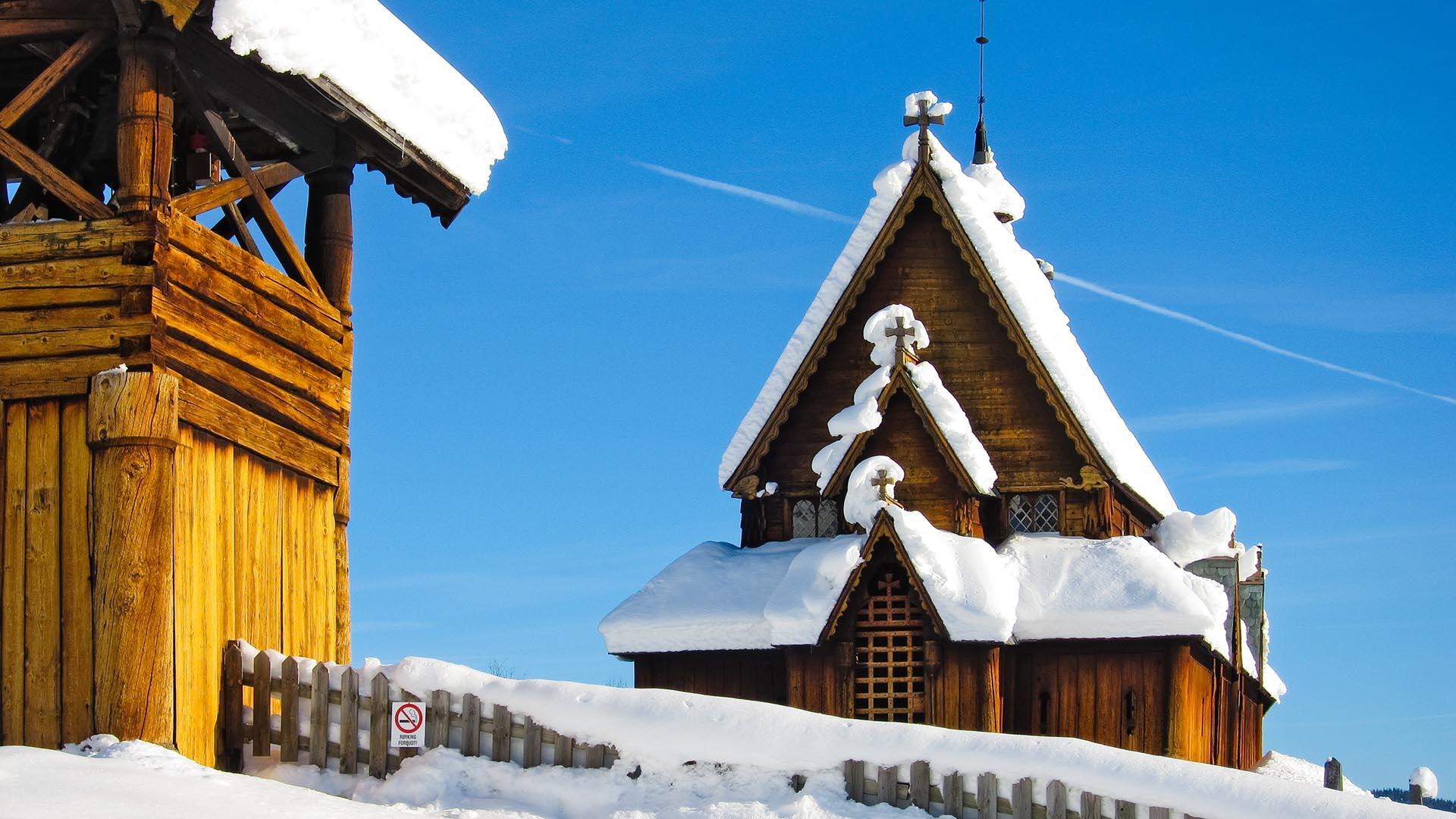 Eine Stabkirche im Schnee gegen einen strahlblauen Himmel. Einen Teil des freistehenden Glockenturms sieht man im Vordergrund.