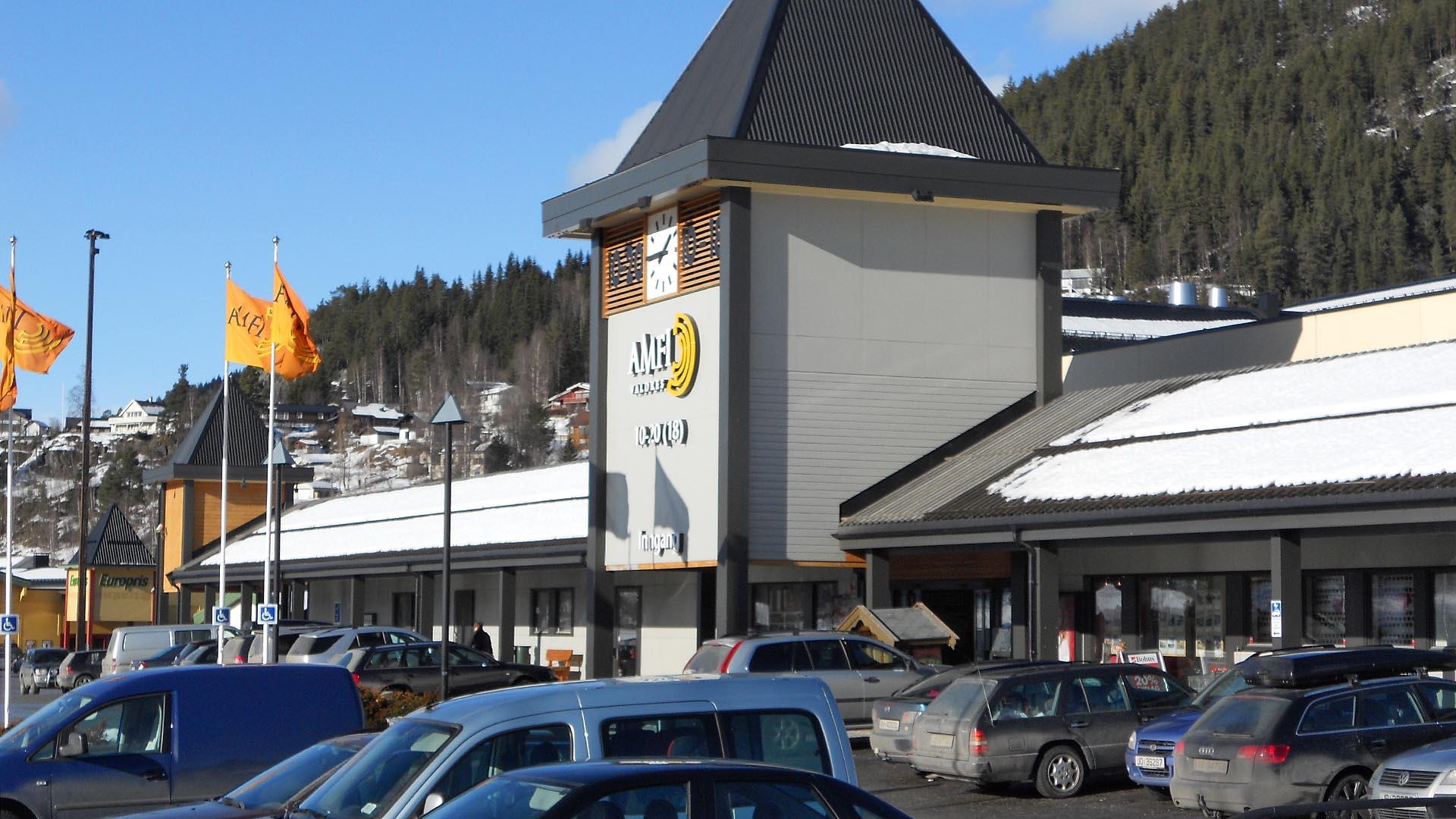 Utsiden av et kjøpesenter med oransje kjøpesenterflagg rett til venstre for det gråmalte inngangspartiet som er formet som et klokketårn.