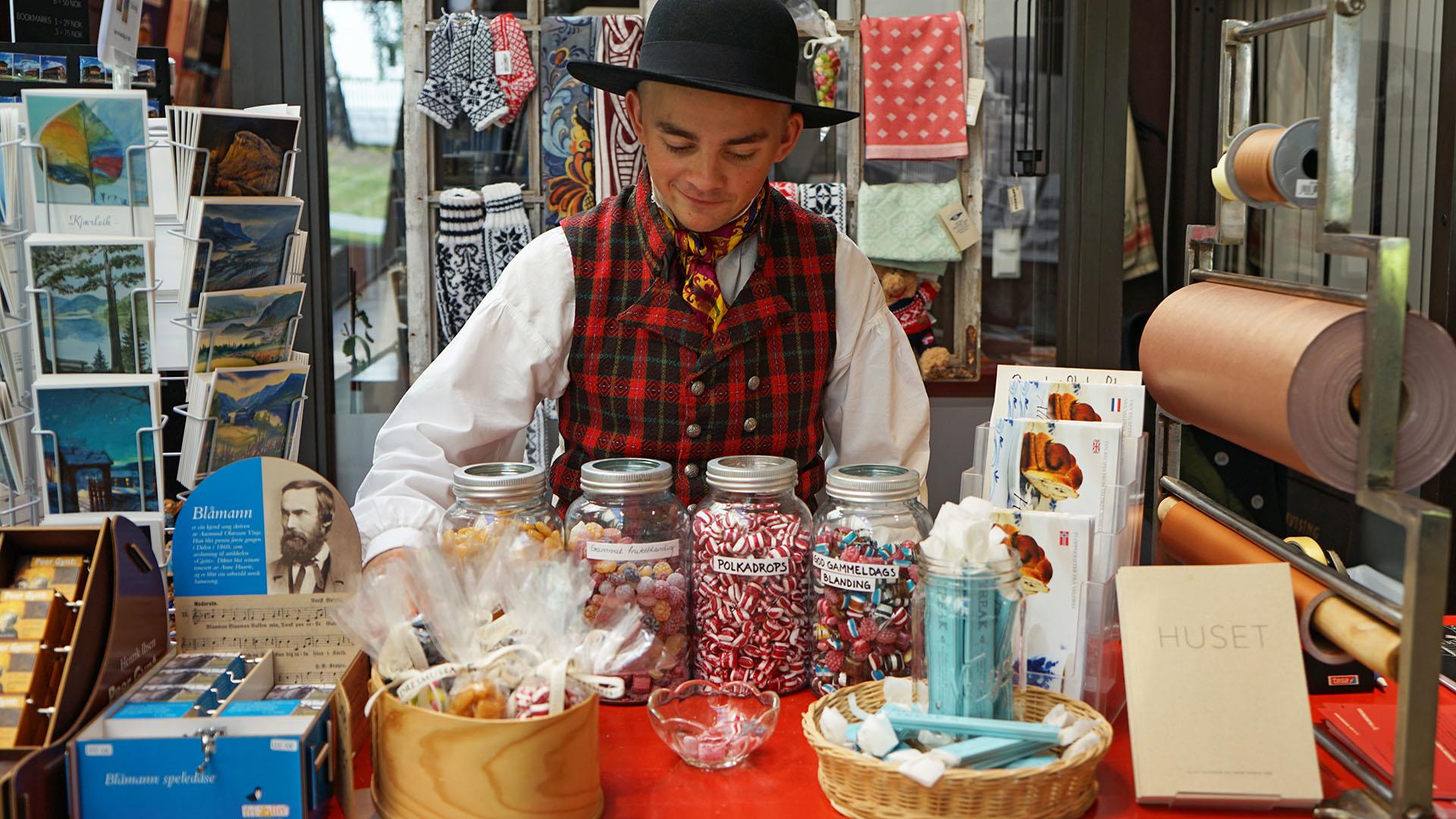 En bunadkledd ung mann bak disken på en museumsbutikk med gammeldagse drops på glass, strikkede votter, kunstkort og bøker.