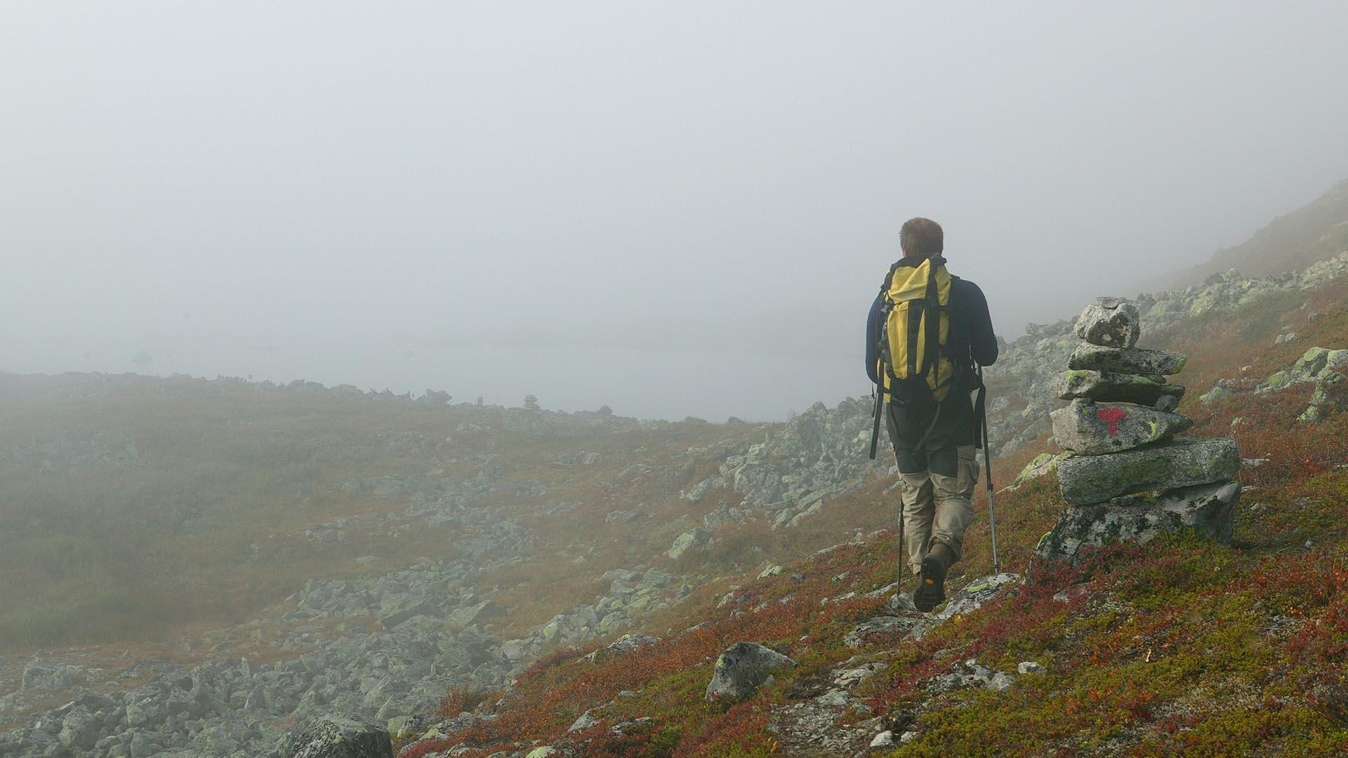 A mountain hiker with a yellow backpack walks past a marking cairn along a trail during fog.