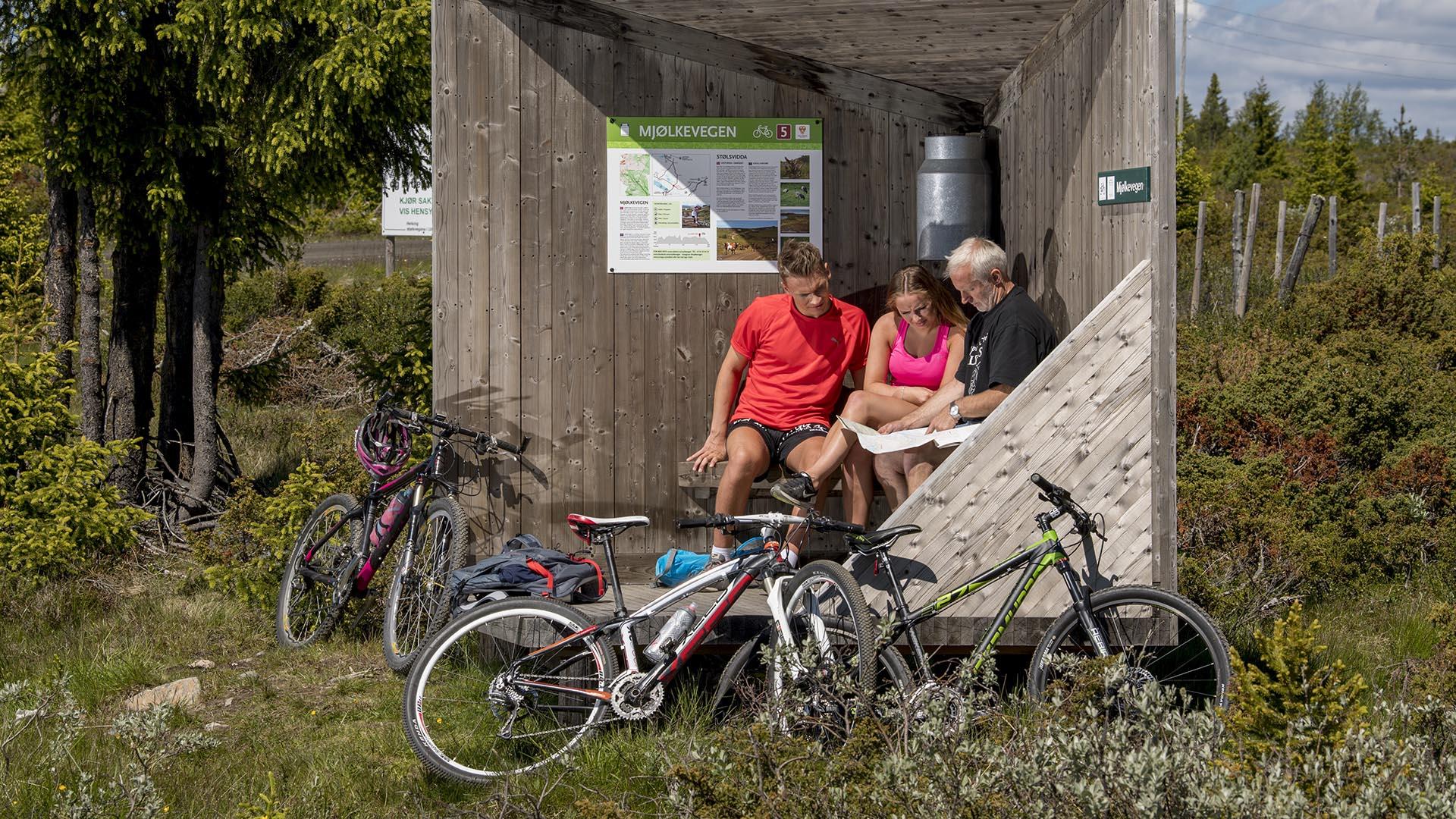 Drei Personen sitzen in einem Designunterstand entlang der Fahrradroute Mjølkevegen.