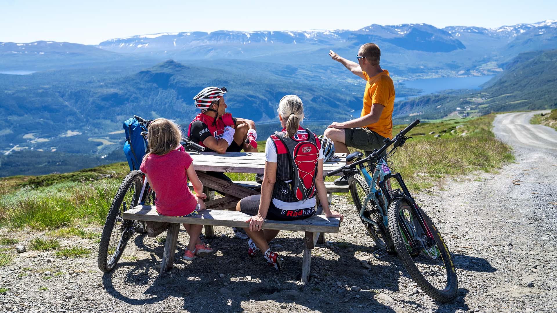 Radfahrer an einem Rastplatz mit Tisch und Bänken an einer Schotterstraße über einen Bergpass mit fantastischer Aussicht ins Tal und zu fernen Bergen an einem schönen und warmen Sommertag.