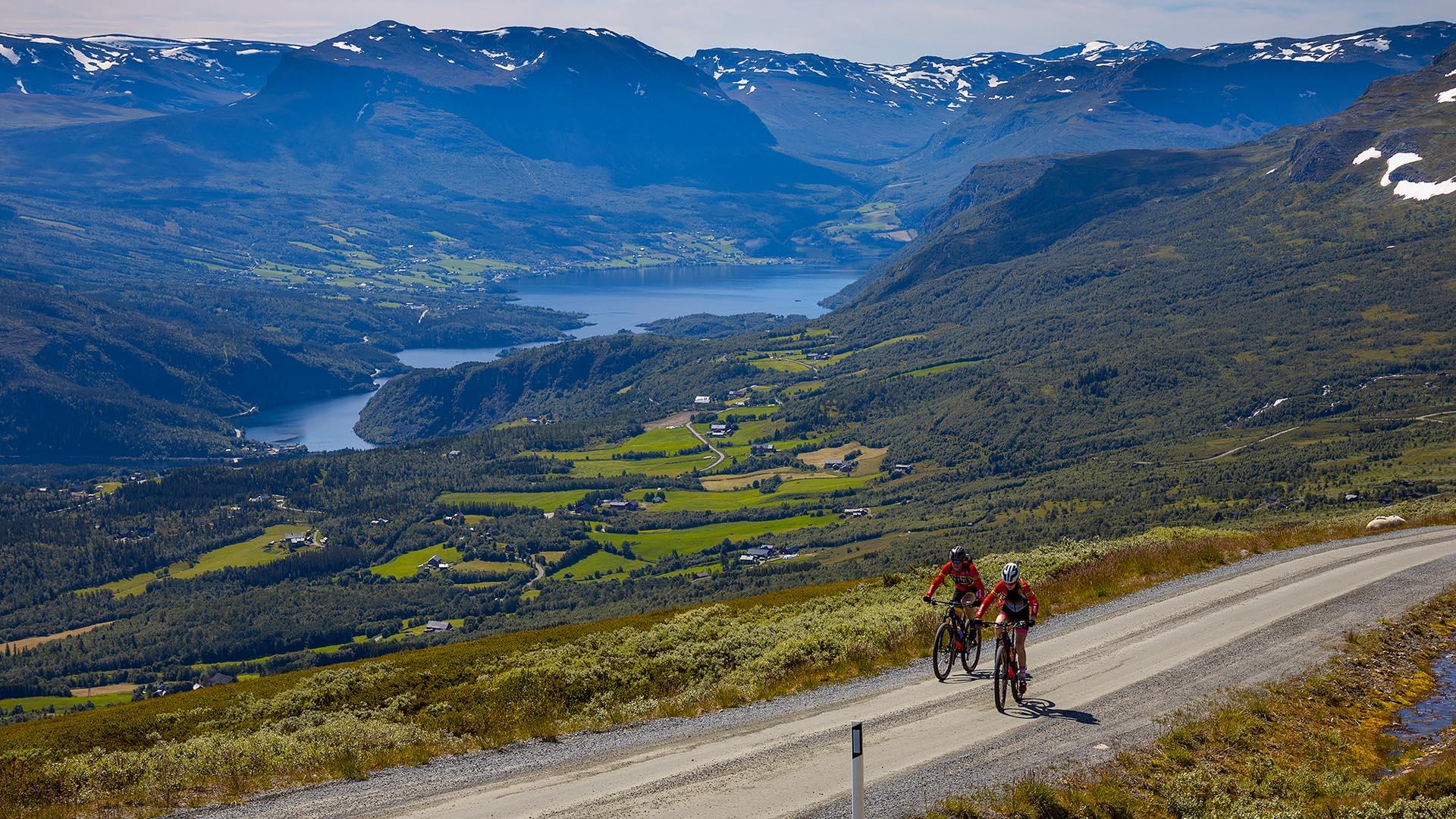 Blikk ovenfra ned på en grusvei med to syklister og en fantastisk utsikt oppover en dal med vann og grønne jorder og fjell en sommerdag.