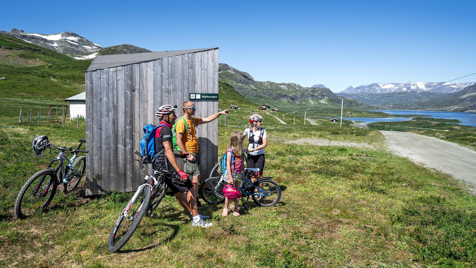 Cyclists taking a break at a rest shelter along a farm road in the mountains with view to a lake and high peaks.