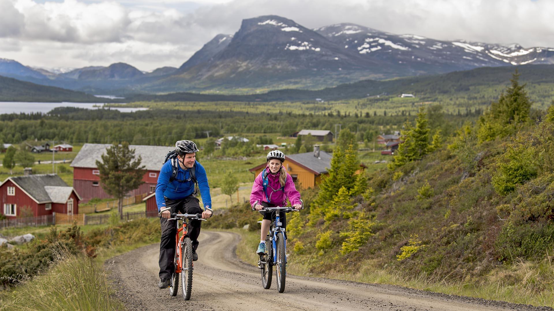 To syklister sykler gjennom et stølslag på en svingete vei. I bakgrunnen ses mektige fjell.