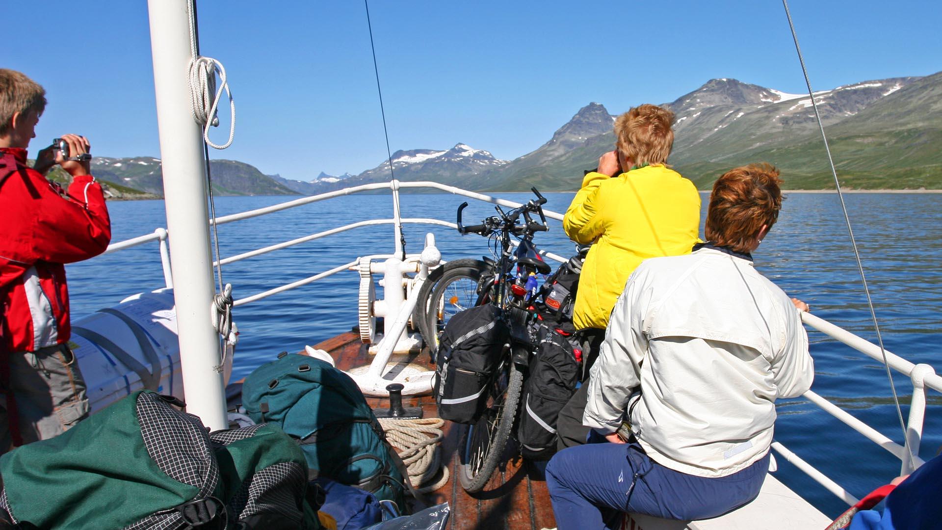 I baugen på en båt på et stort fjellvann med totusenmetertopper langs bredden. Folk sitter og nyter utsikten en klar sommerdag, og noen har med sykkel ombord.