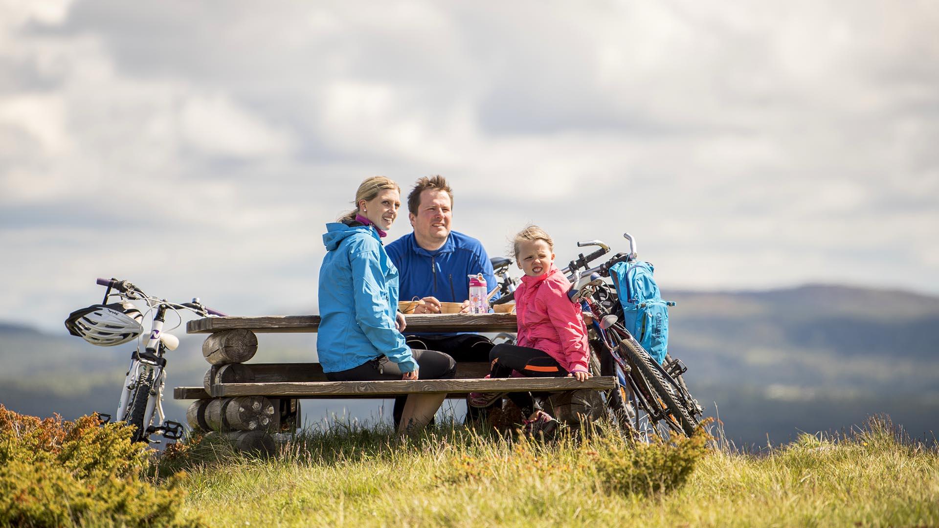 A family has a break on a cycling trip at a resting area with table and benches in the mountains.