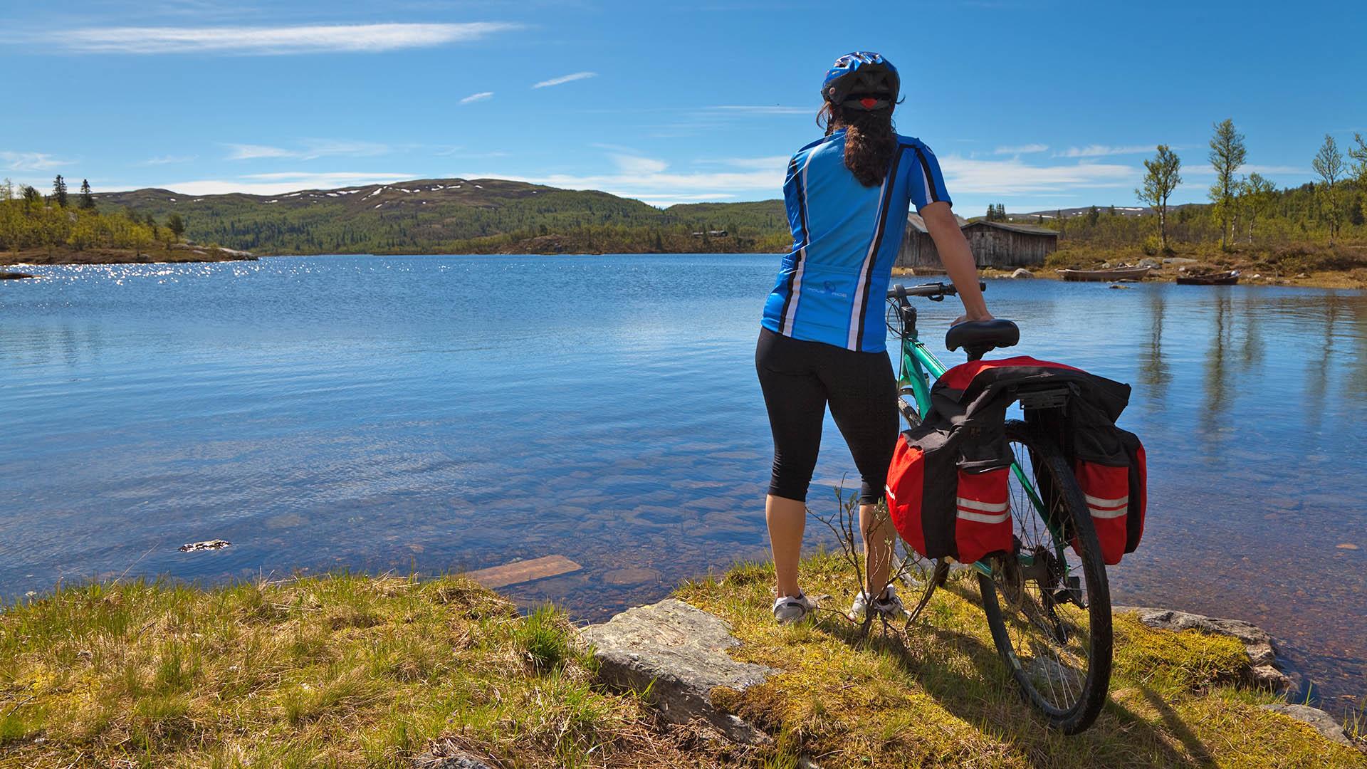 A female biker in cycling outfit is standing next to her bike on the shore of a mountain lake.