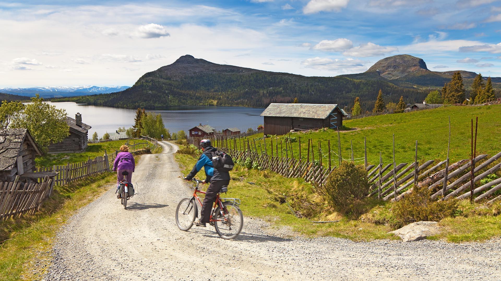 Two cyclists cycle on a gravel road that leads downhill towards a lake in beautiful surroundings with green mountain meadows, a cabin and mountains behind the lake.