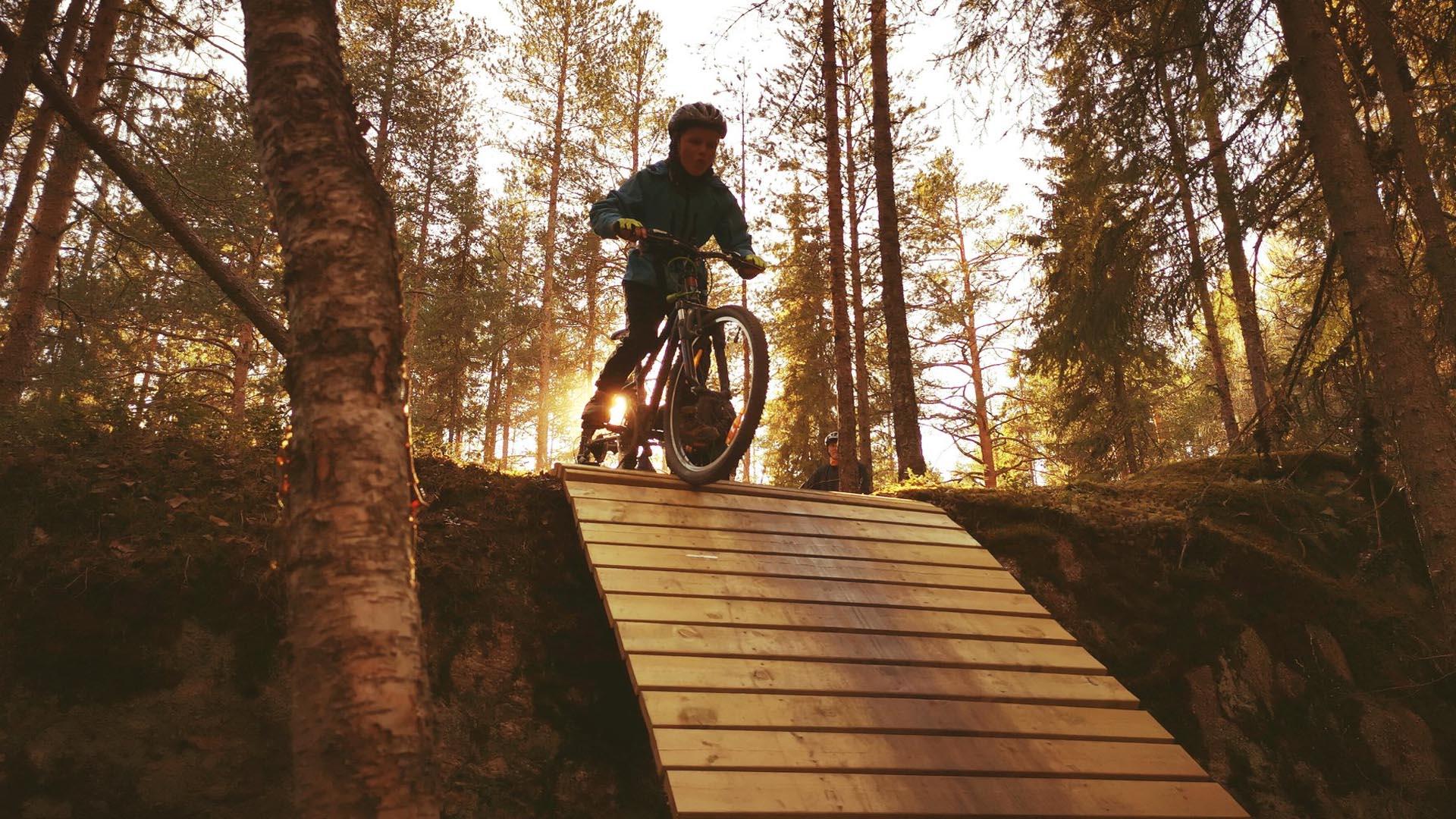 En gutt på terrengsykkel på toppen av en hellende trebro i skogen i motlys.