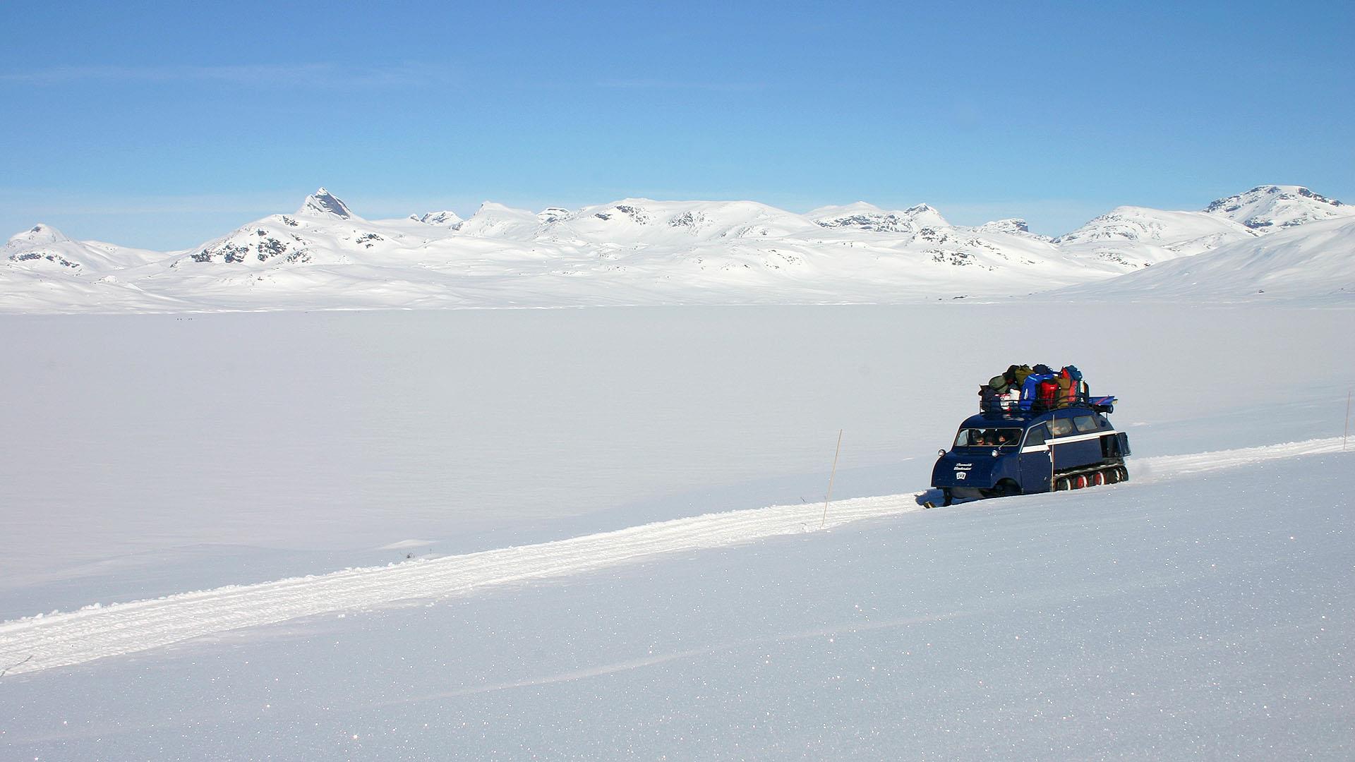 Beltebil kjører på snø en skyfri vinterdag med Jotunheimen i bakgrunnen.