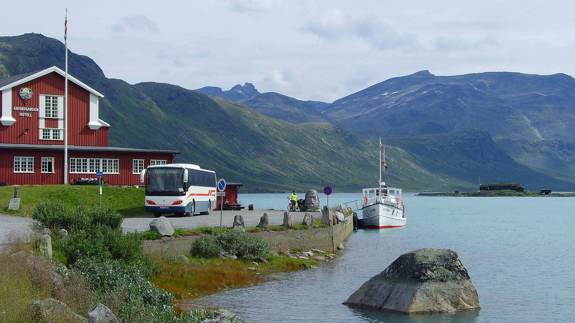 A historic passenger boat on a turquoise mountain lake, a bus on the quay, a red wooden hotel and several twothousand-meter-summits in the background.
