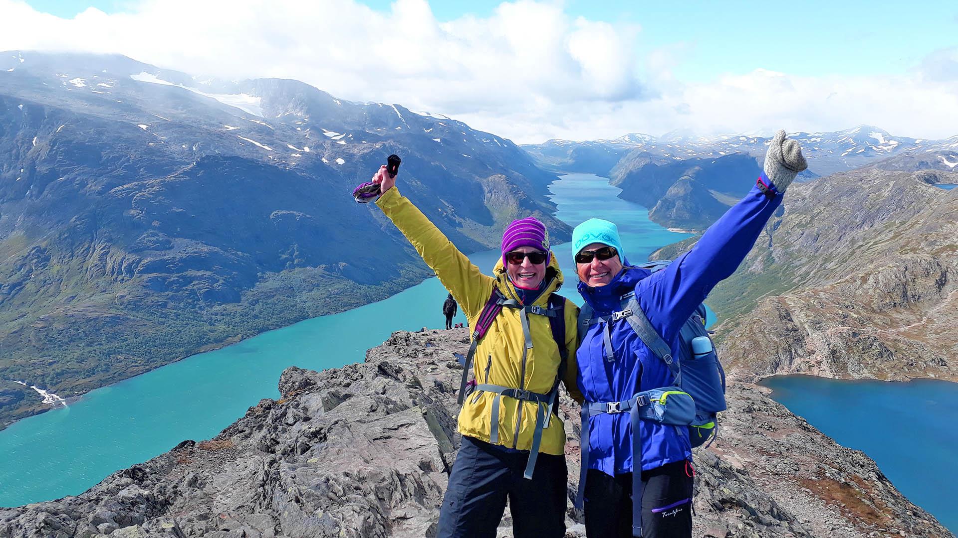 Two hikers in outdoor clothes with their arms in the air are standing on a narrow ridge between two lakes in the high mountains.
