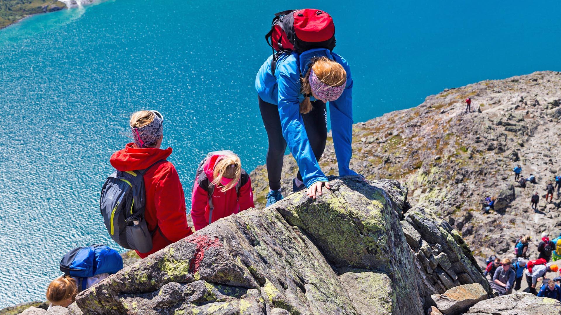 Children scramble opp a slightly exposed section of a rocky ridge in the mountains that lies above a green-blue lake.