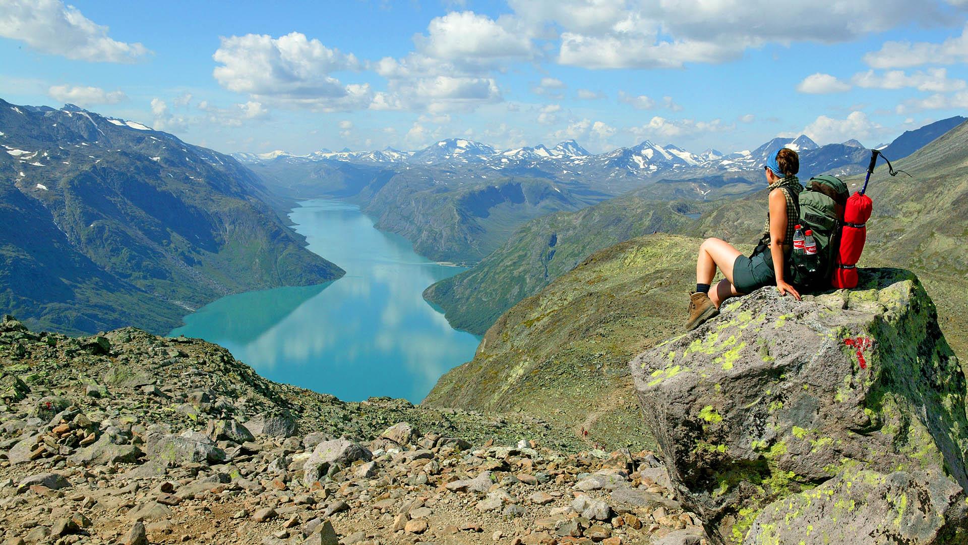 En kvinne sitter på en stor steinblokk og ser innover fjellet med et stort vann omkranset av høye fjell en fin og varm sommerdag.