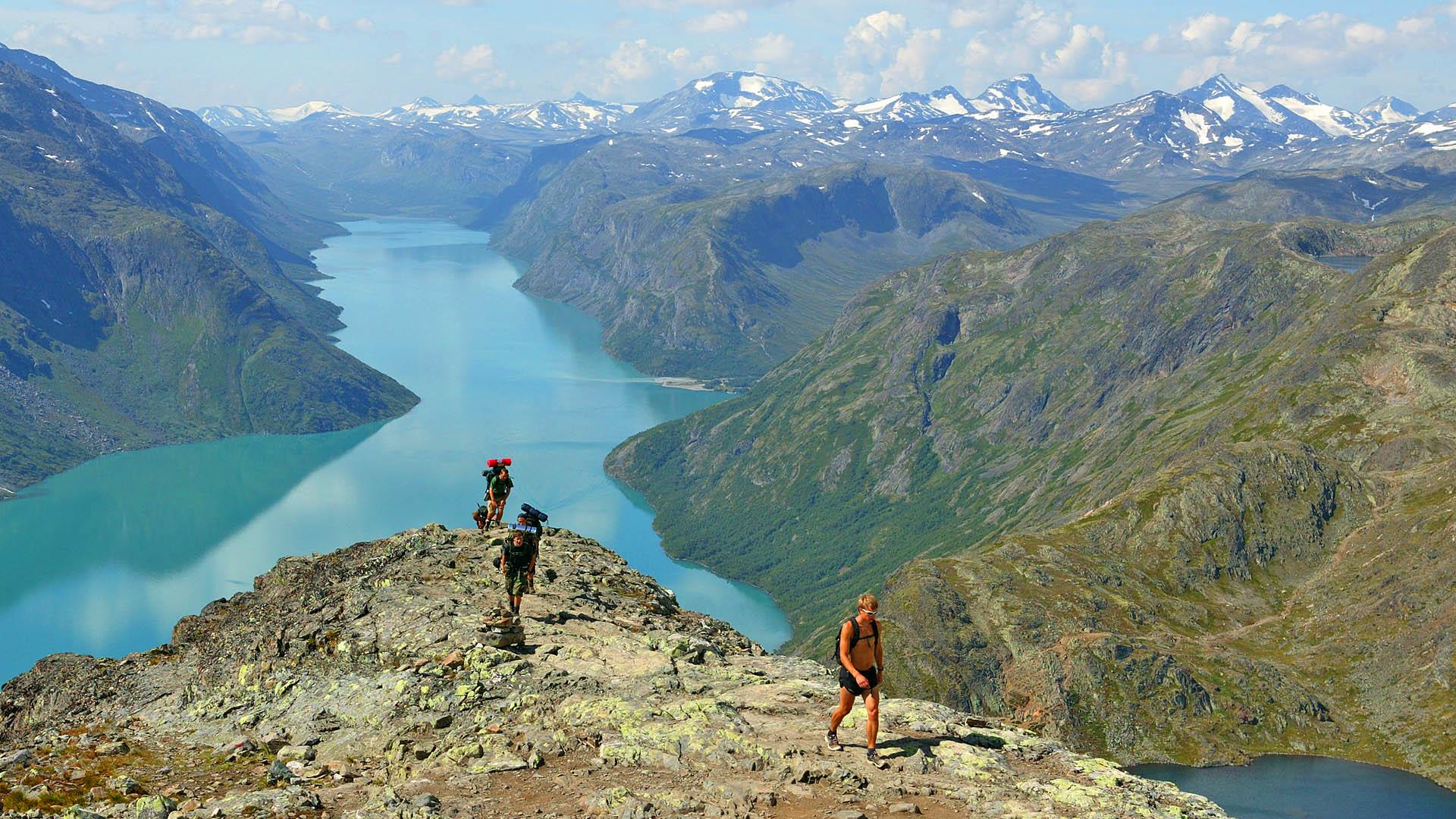 Hikers emerge over a ridge edge with a long and narrow mountain lake surrounded by high peaks in the background on a fine and warm summer day.