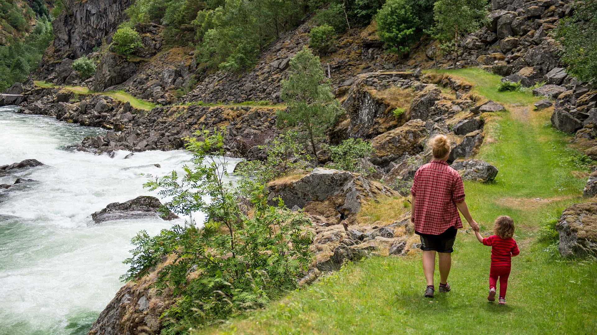 A mother and a small child walk on a historical road with grass cover along a river in a gorge.