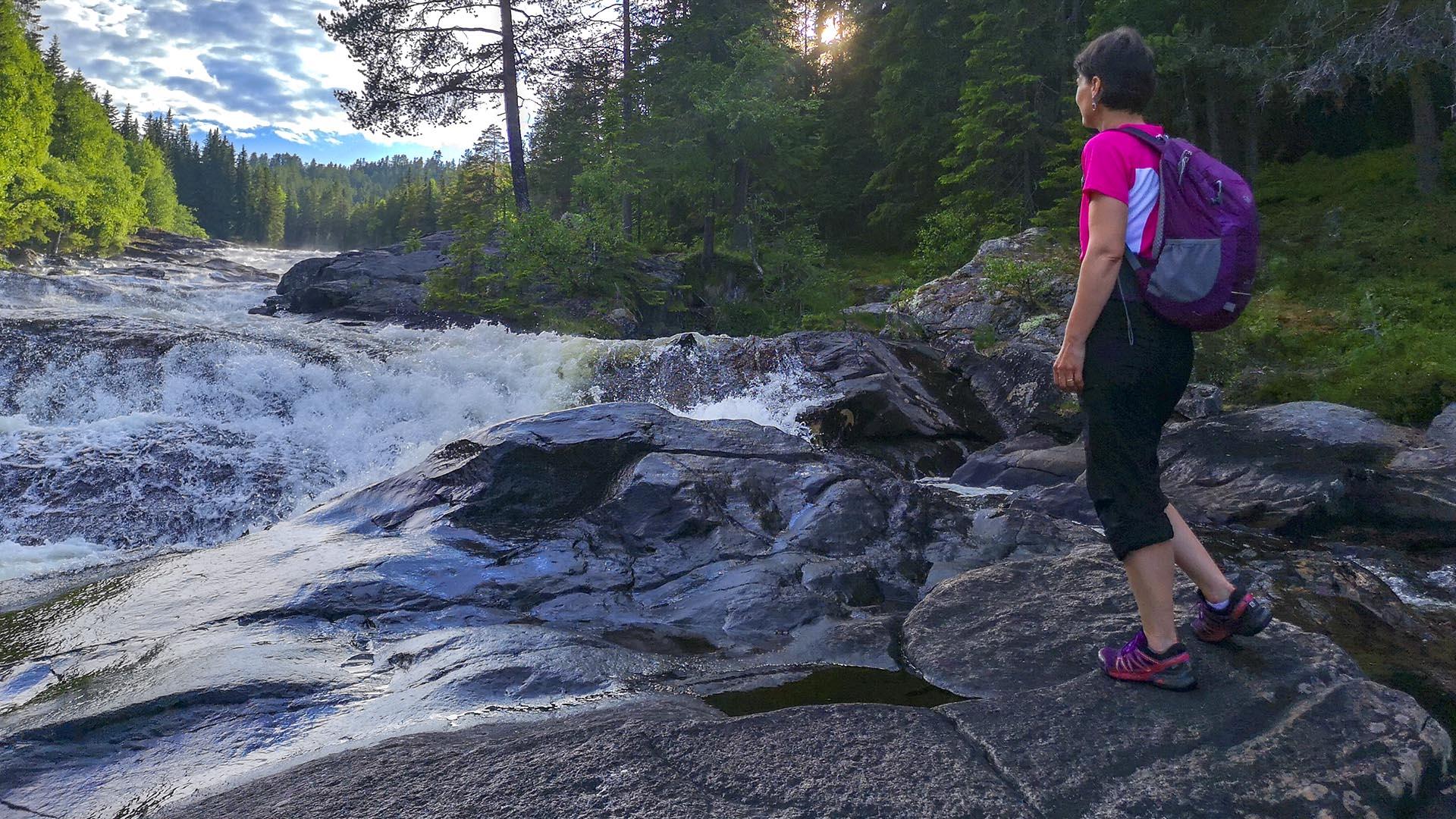 A woman is standing on flat rocks by a river with fast rapids in a forest