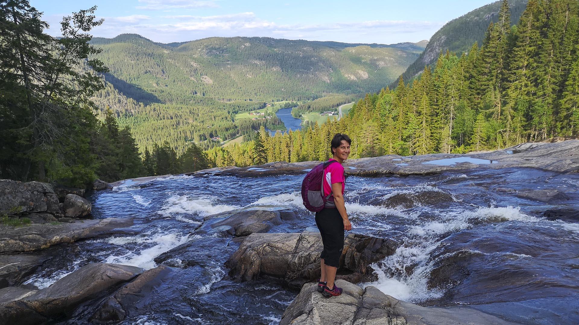 A woman with a day pack isn tanding on a rock in a river that drops and disapears into a waterfall behind her. In the background, far below, a view into the main valley with a large river and forests.
