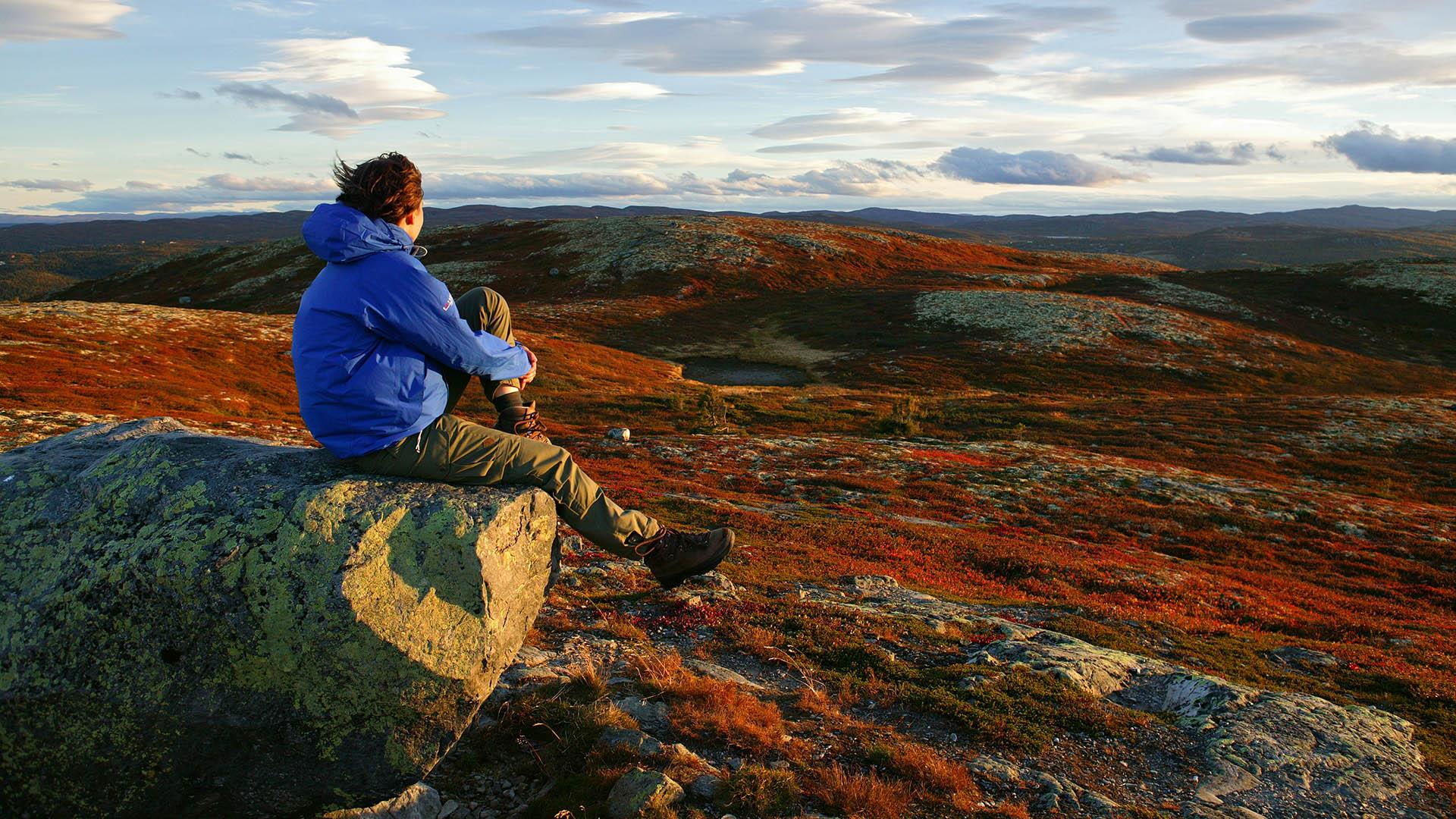 Kvinne sitter på en stein på en slak fjelltopp en høstdag med rød lyng på slettene i bakgrunnen.