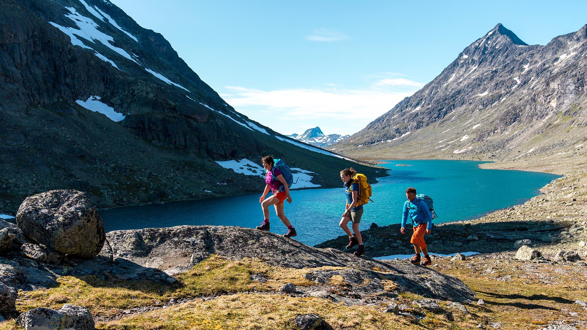 Tre sommerkledde vandere med ryggsekk i en dal i høyfjellet med et vann og spisse, høye fjell omkring.