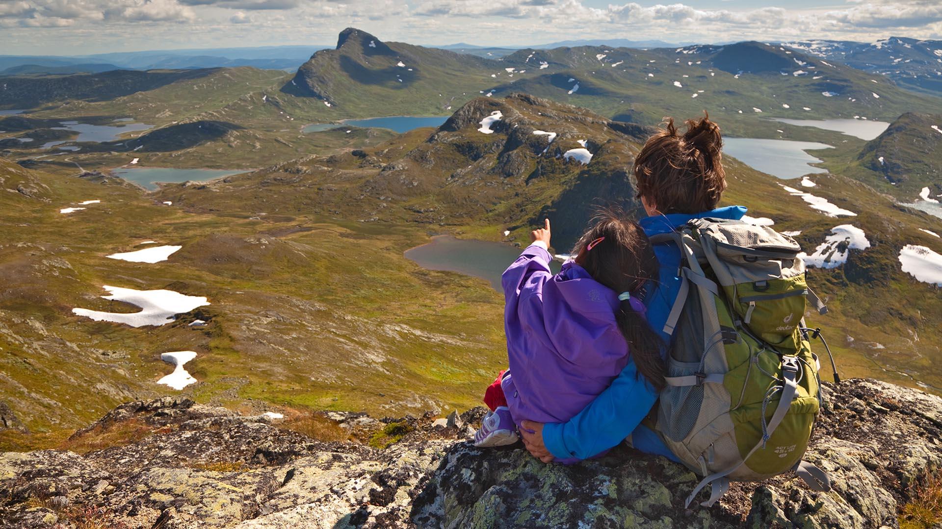 Kvinne og barn sitter på fjelltopp og nyter utsikten mot fjell og innsjøer.