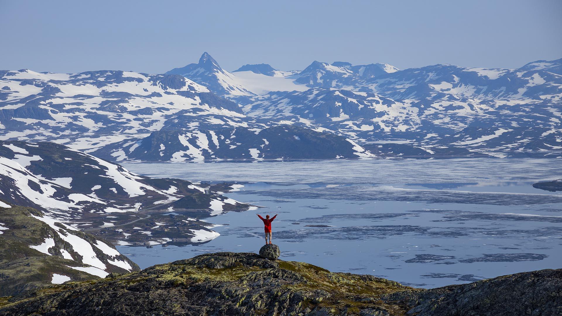 Far away a person stands on top of a rock enjoying view over a lake and snowspotted mountains in the distance.