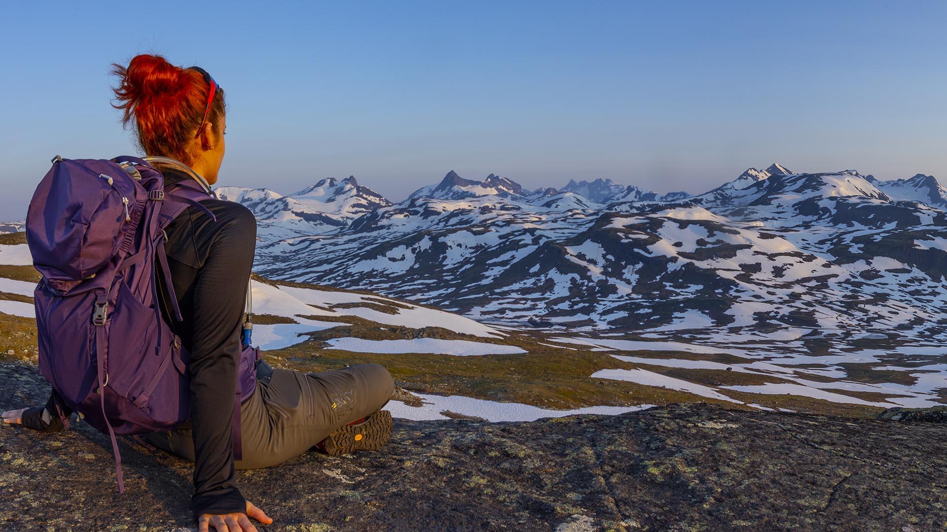 Eine Frau sitzt im Vordergrund und genießt die Aussicht zum Hochgebirge mit vielen Schneefeldern im lila Morgenlicht.