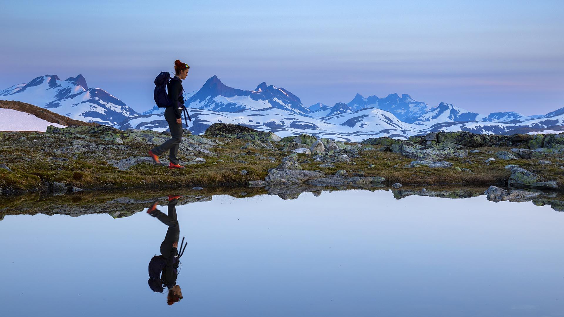 Eine Frau wandert an einem glasklaren Wasser vorbei und spiegelt sich darin. Hohe, spitze Berge im Hintergrund. Lila Morgenlicht vor Sonnenaufgang.