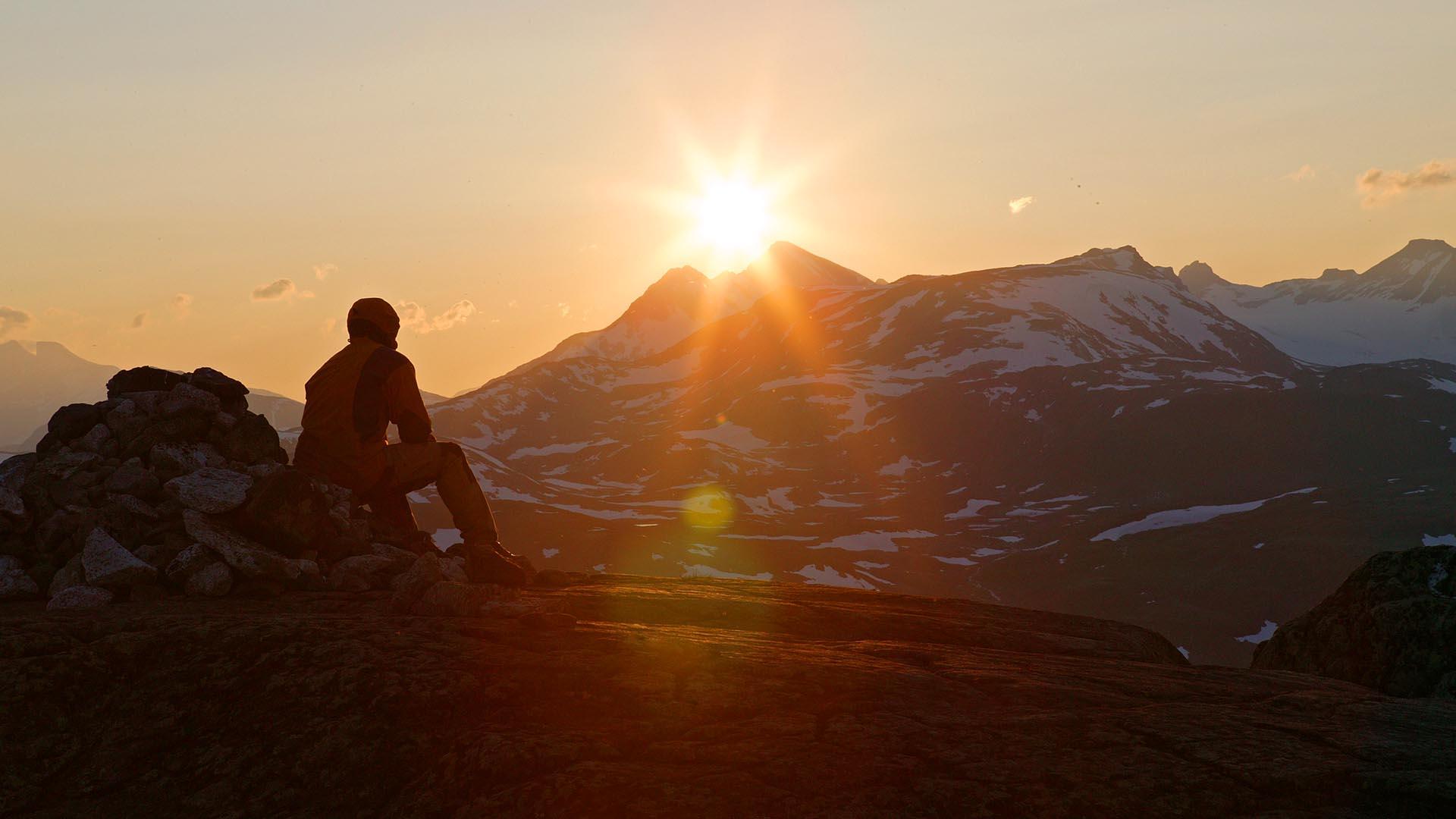 Mann sitter på fjelltopp og ser solen gå ned bak et annet fjell.