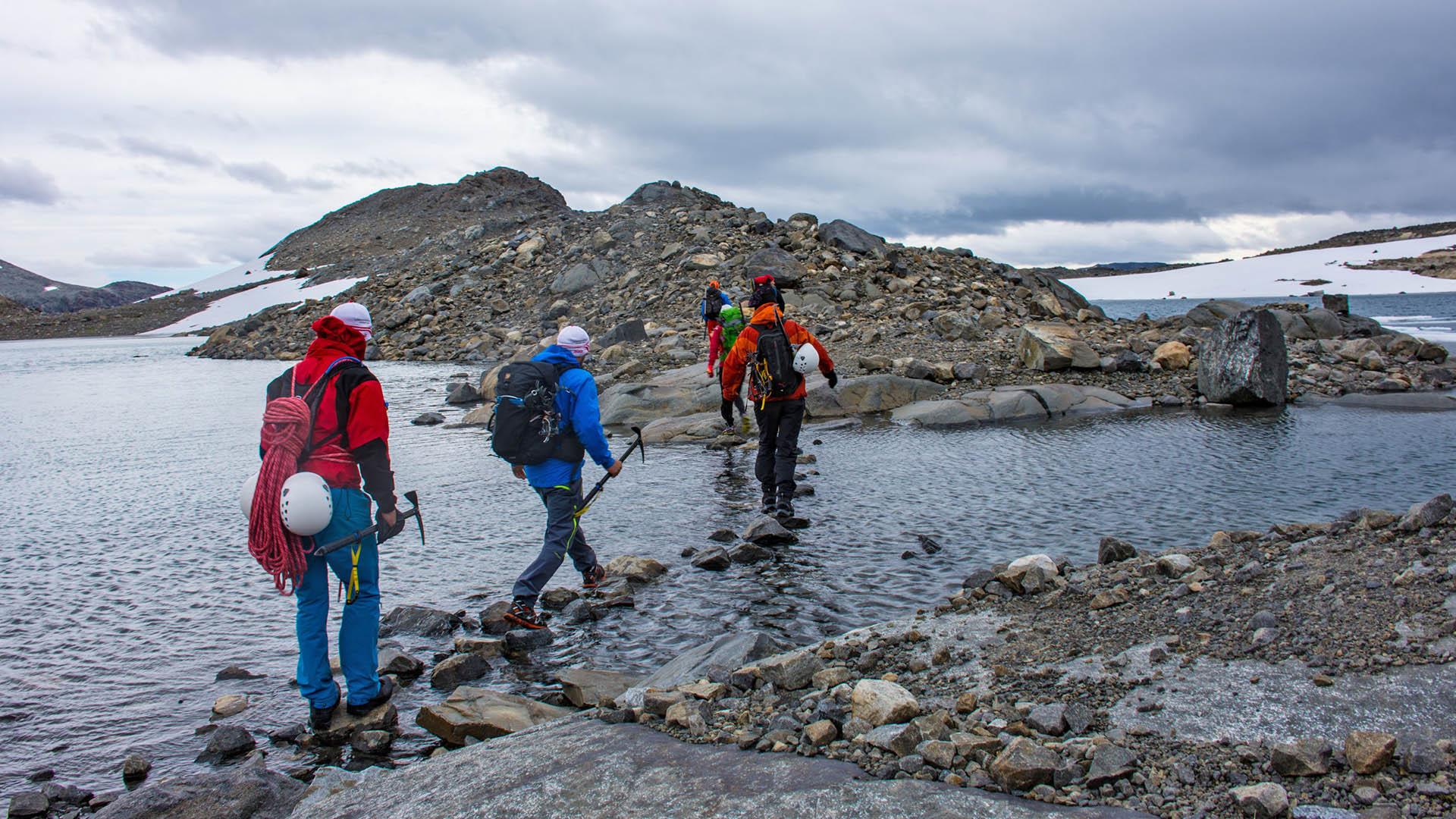 Bergsteiger mit Rucksäcken, Helmen und Eispickeln überqueren einen Fluss in einer Stein- und Schneeöde im Hochgebirge.