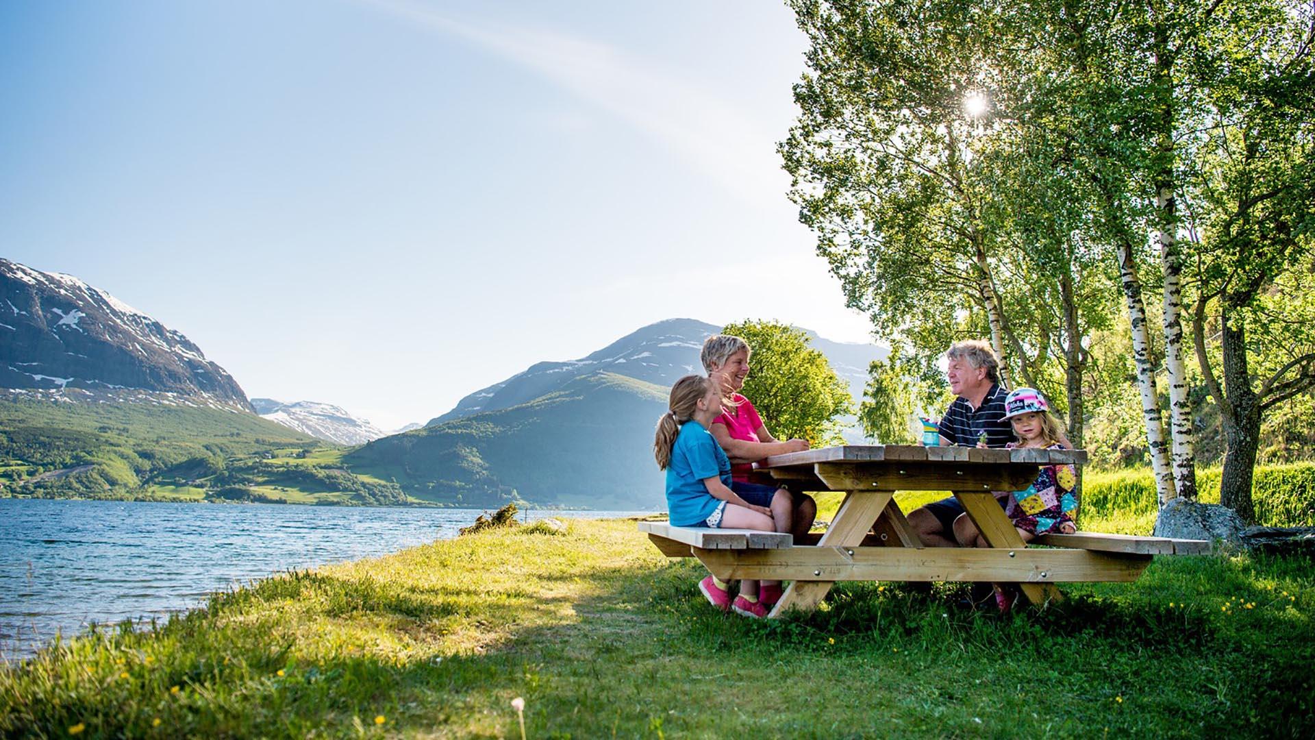 A family sits at a wooden table with benches on a grass-overgrown rest area by a lake under a birch tree. In the background mountains rise.