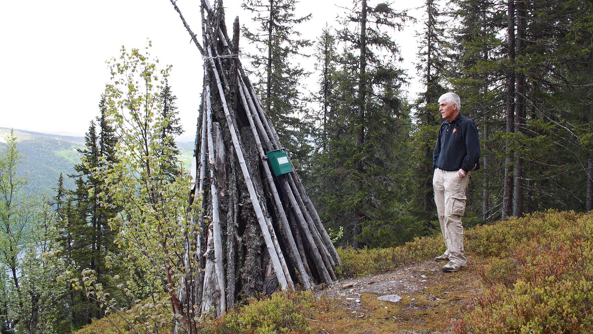 A man is standing next to a historical wooden beacon which is placed on an outcrop in a forested hillside.