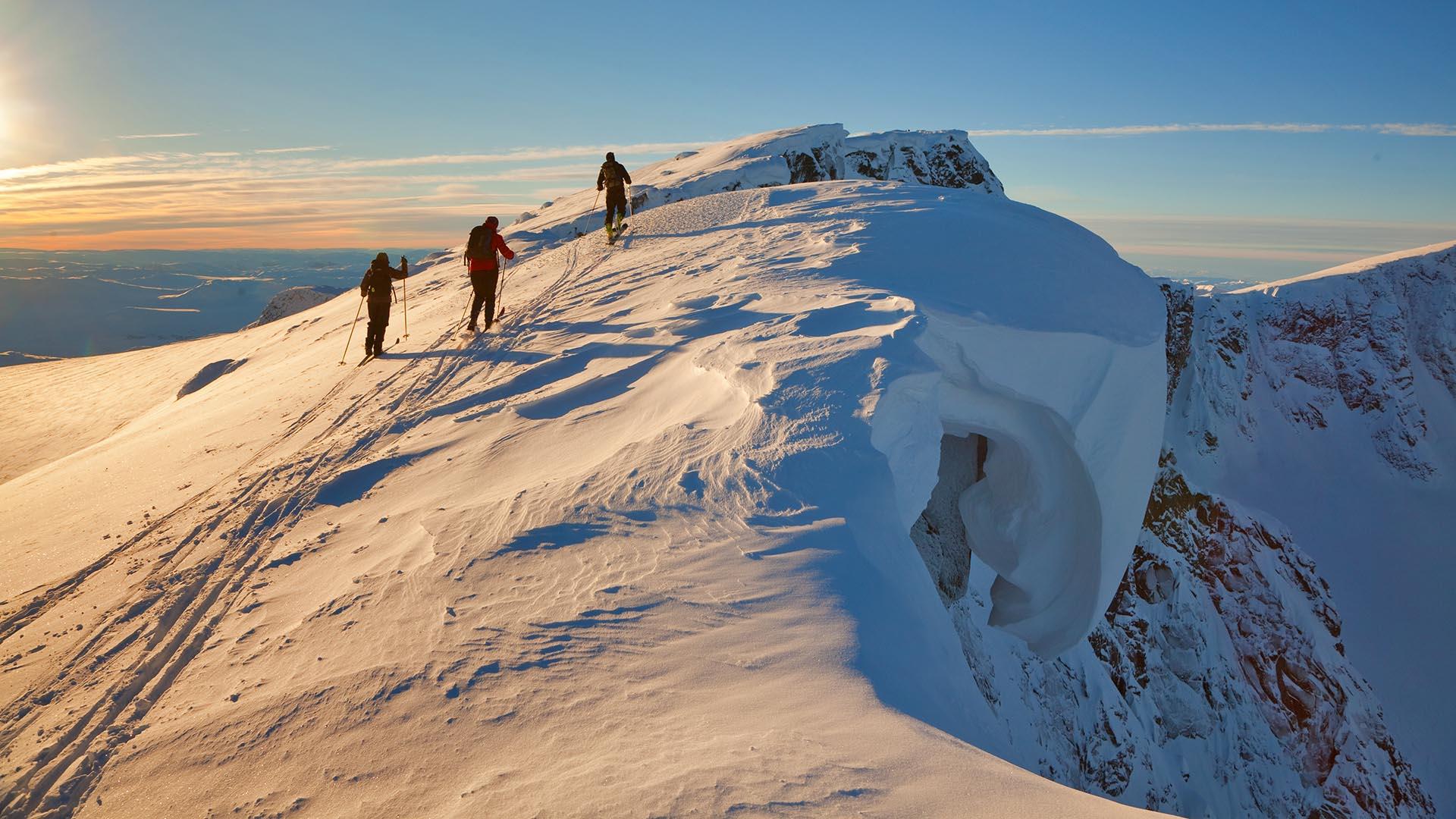 3 personer på vei opp en fjelltopp på fjellski. Blå himmel i bakgrunnen.