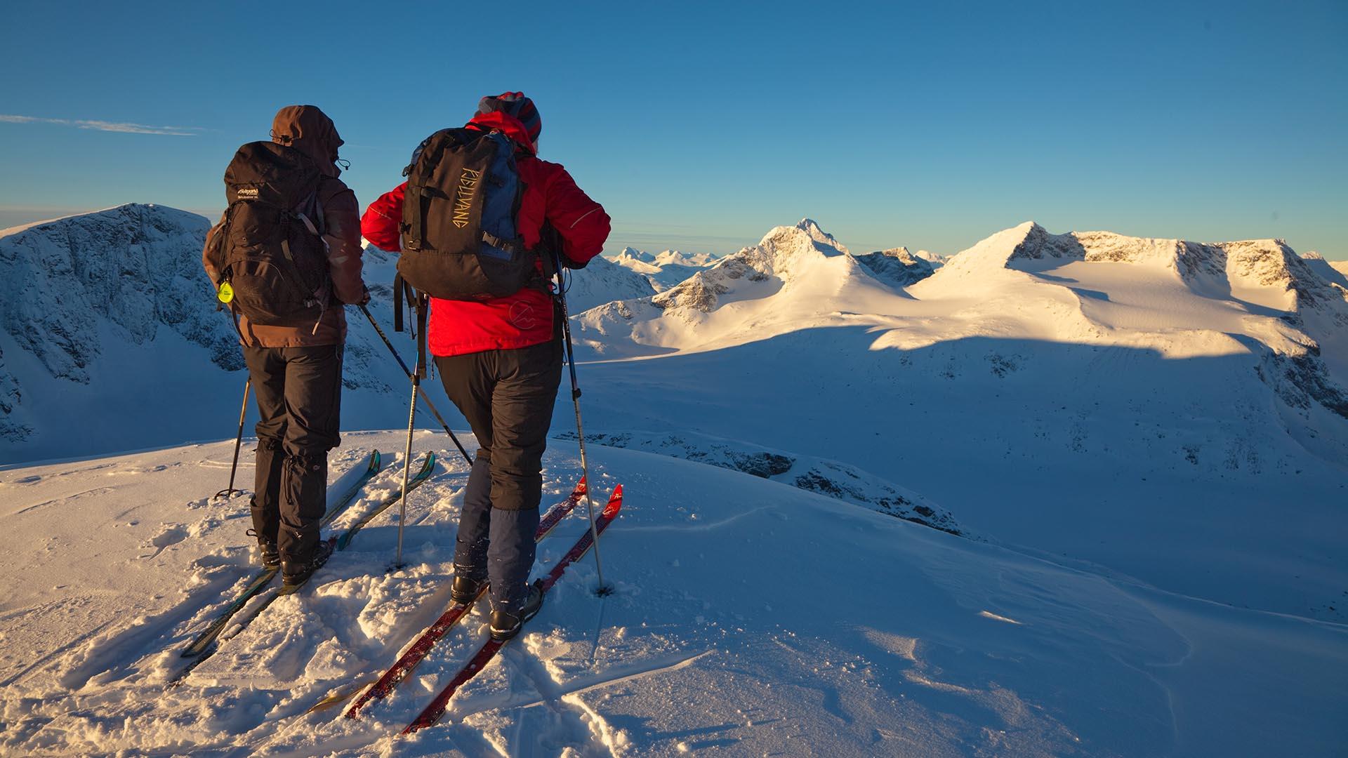 Two persons on skis taking a breather on a mountain top looking towards a snow covered mountain landscare a wintersday with no clouds.