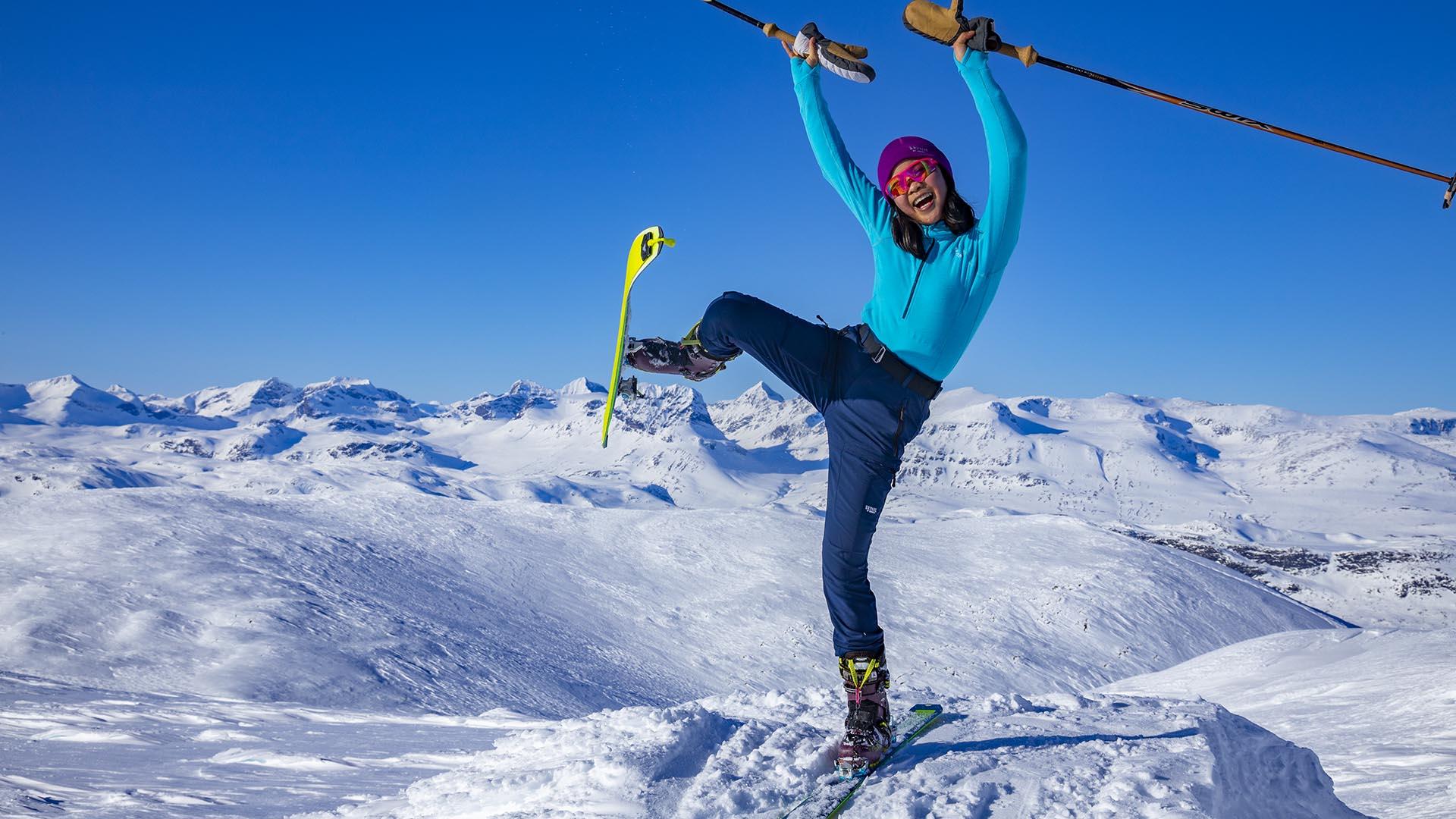 Jente på fjelltopp med randoski, superglad og strekker hender og den ene foten med skien i været. Langstrakt utsikt over snøkledde fjell bak.