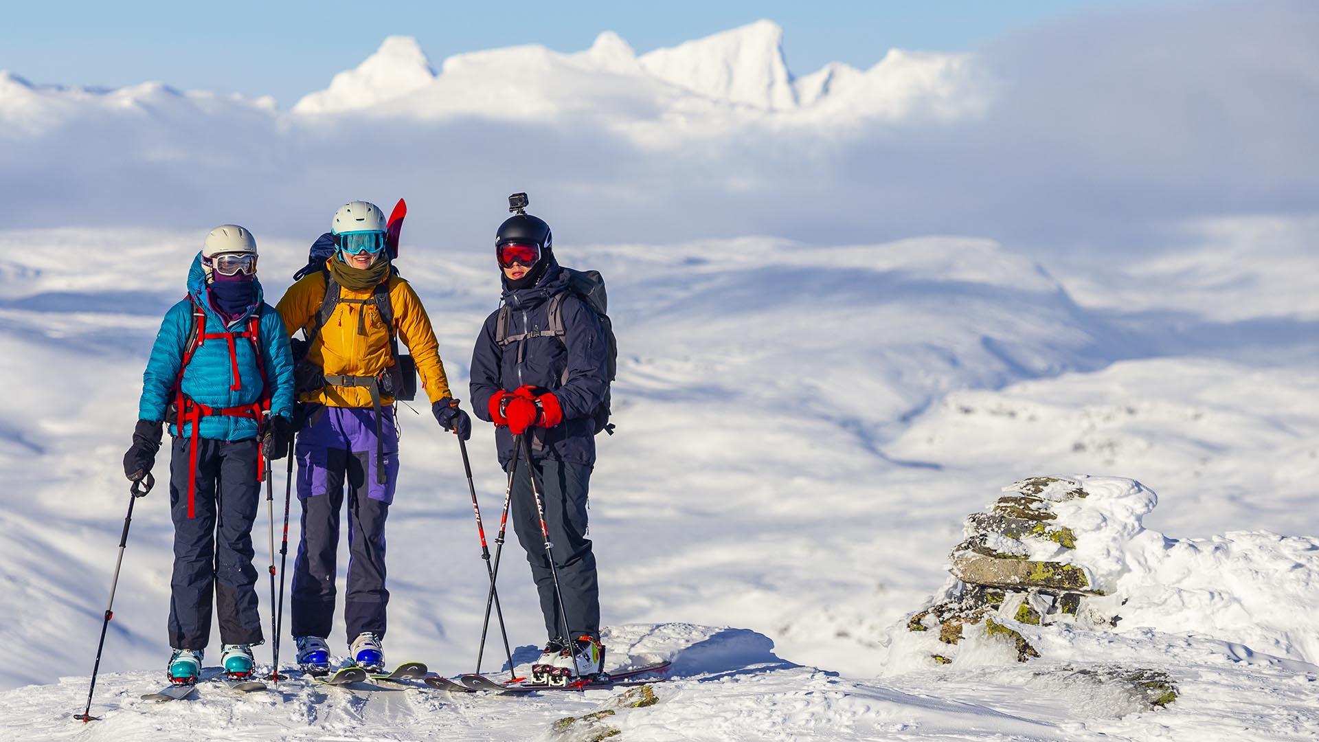 Tre personer på fjelltopp med randoneeski, vid utsikt over snødekt fjellandskap