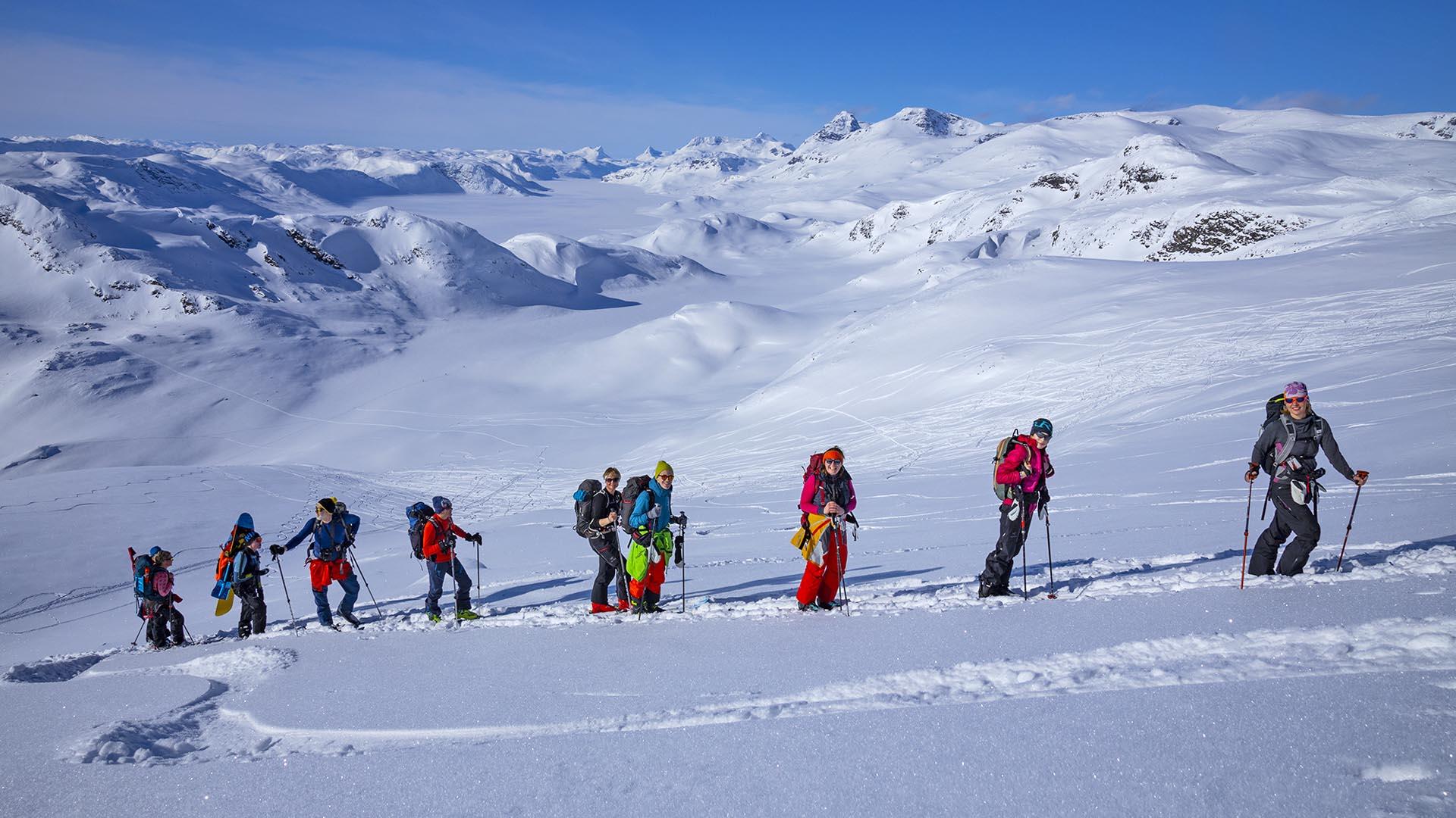 Group of 9 persons on their way up a mountain on randonee skis. Snow covered mountains in the background.
