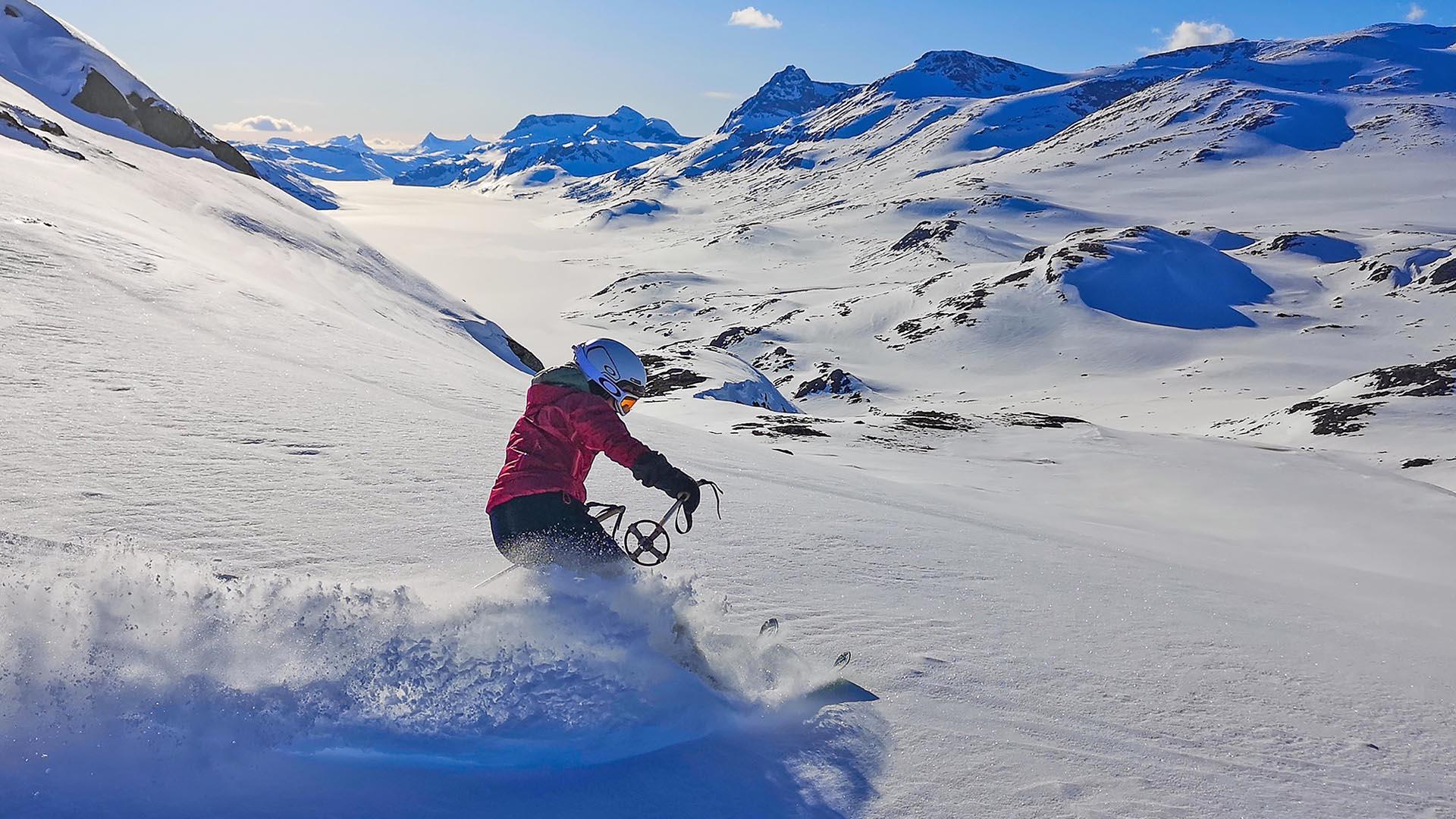 Eine Tourenskifahrerin legt eine Kurve hin, dass der Schnee stiebt. Hochgebirge im Hintergrund.