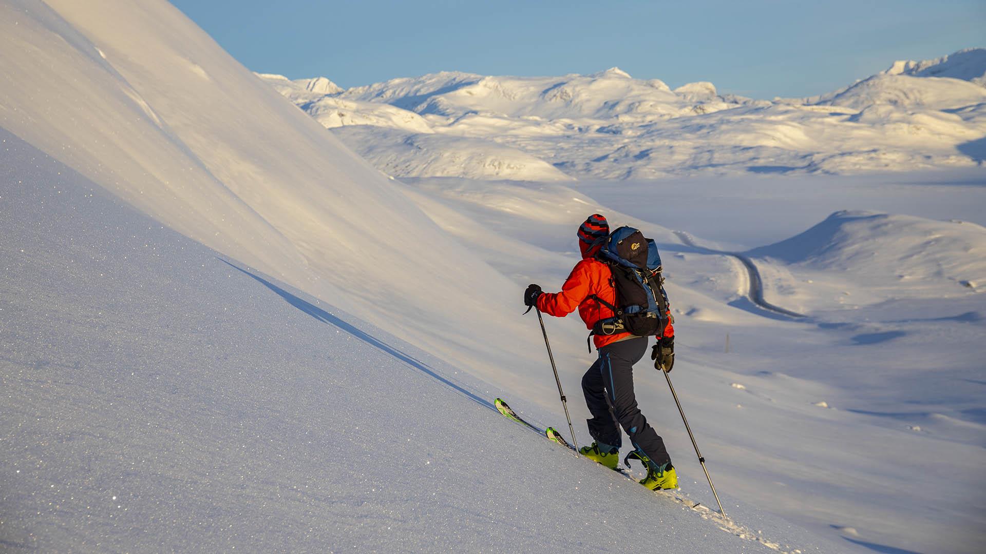 Person på vei opp fjellside med randoneeski, vid utsikt over snødekt fjellandskap