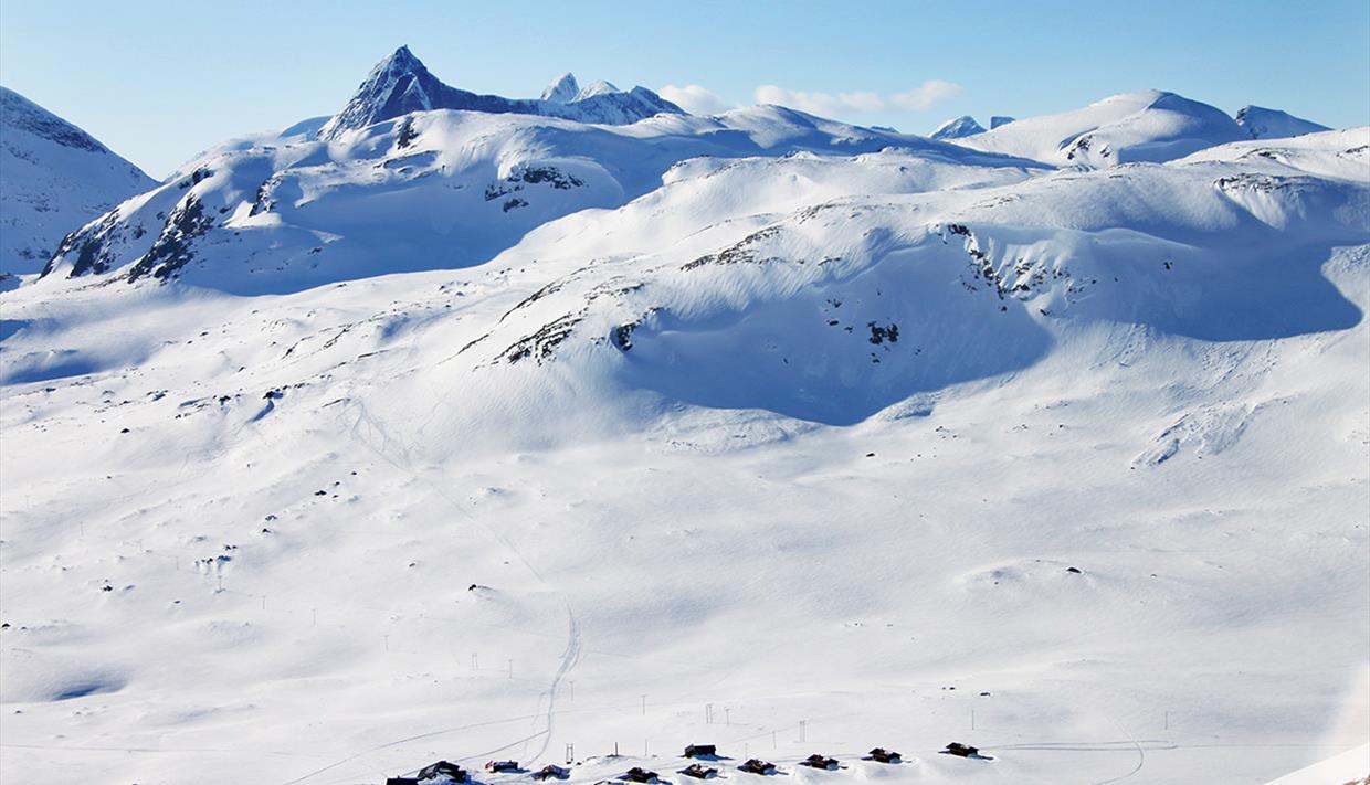En liten fjellstue og et knippe hytter i et nedsnødd høyfjellslandskap fra fugleperspektivet