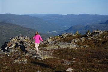 En jente i rosa genser på tur langs en fjellside med vid utsikt bakenfor.
