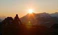 Ein Mann sitzt auf einem Felsen und sieht gen Sonnenuntergang hinter einem hohen Berg.