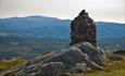 The summit cairn at Jutulen rises proudly on top of a rocky outcrop. Forested hillsides and a higher mountain in the background.